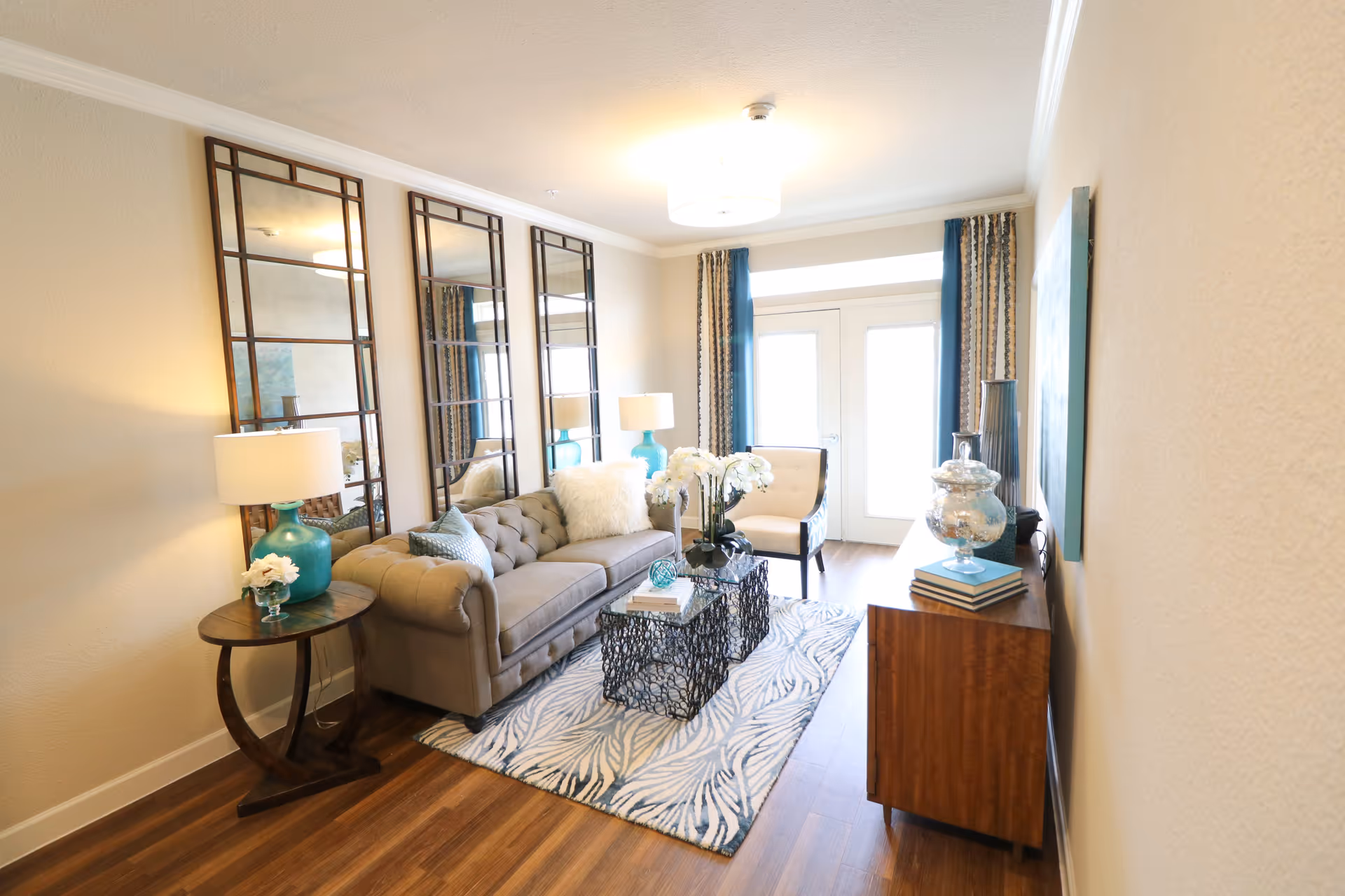 A bright and cozy living room with a beige tufted sofa adorned with pillows, a white armchair, and a patterned area rug. The room features three tall decorative mirrors on one wall, two turquoise table lamps on side tables, and a wooden cabinet with decorative items. French doors with curtains allow natural light to fill the space.