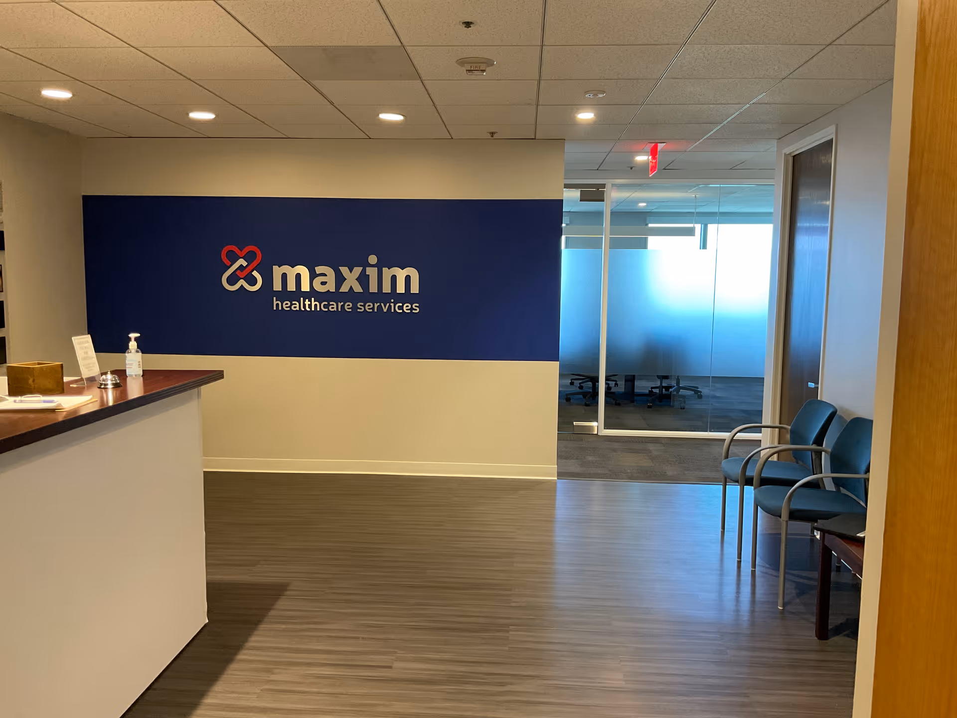 Reception area with blue Maxim Healthcare sign, front desk on left, glass doors, blue chairs.