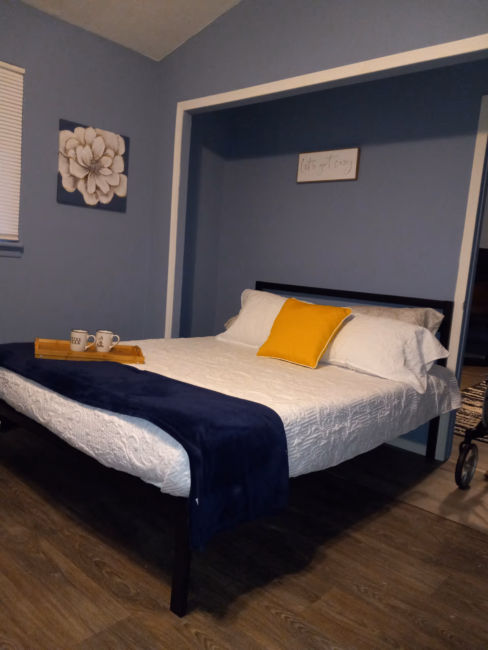 A bedroom with blue walls, white bedding, navy throw, yellow pillow, and a walker nearby.