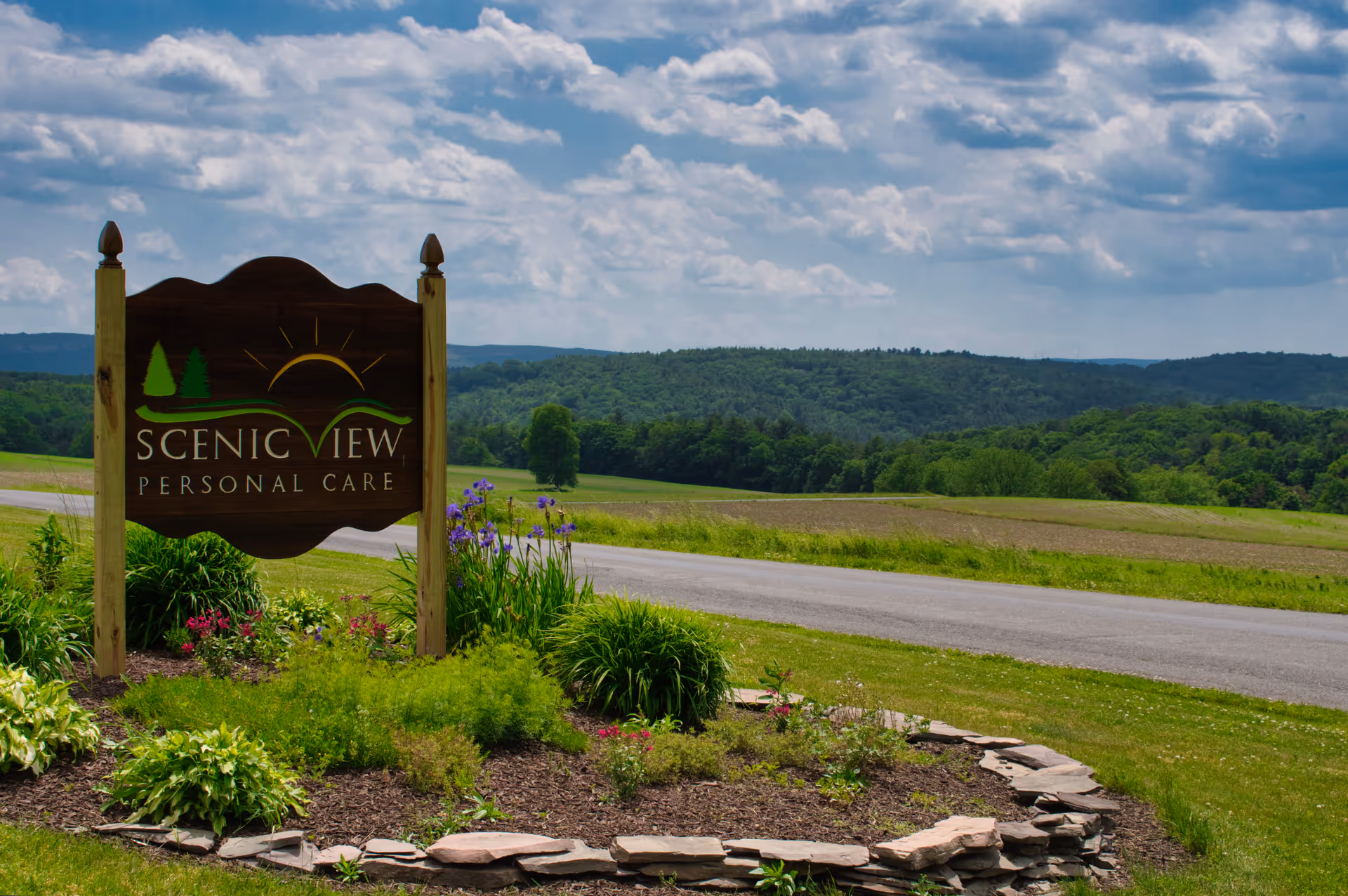 A wooden sign reading Scenic View Personal Care near a grassy roadside with hills.