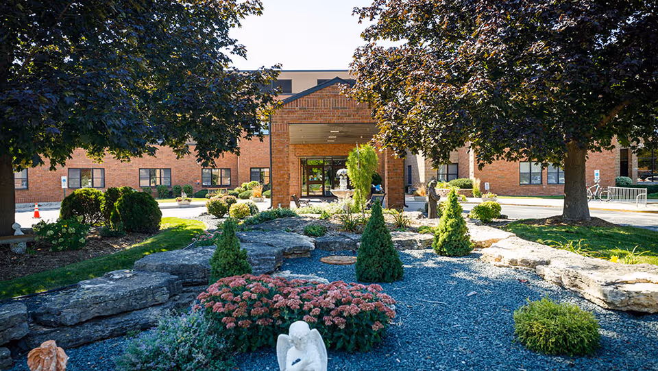 Brick senior living facility entrance with landscaped garden and trees.