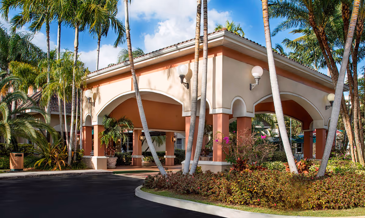 Exterior view of a building entrance with a covered driveway supported by columns, surrounded by palm trees and landscaped bushes under a partly cloudy sky.
