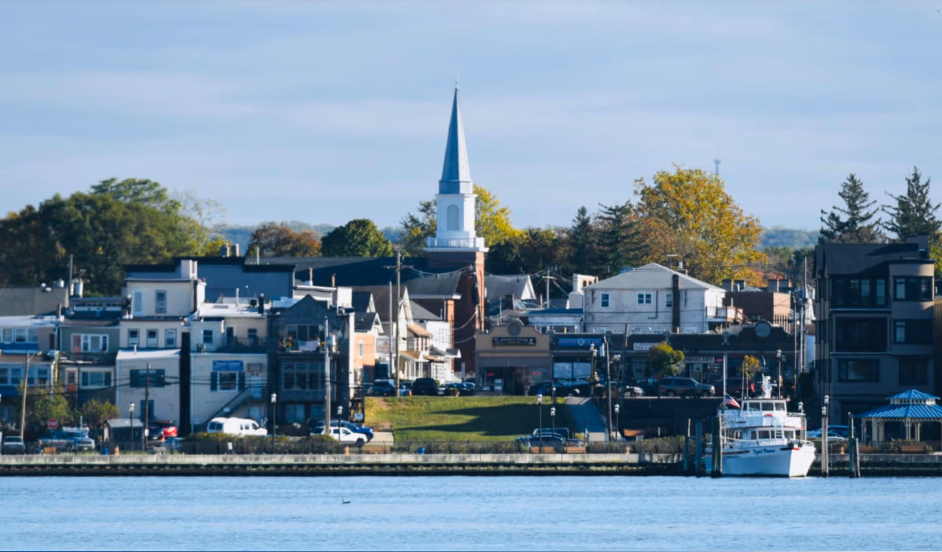 Waterfront town with church steeple, row houses, trees, and a docked boat along the shore.
