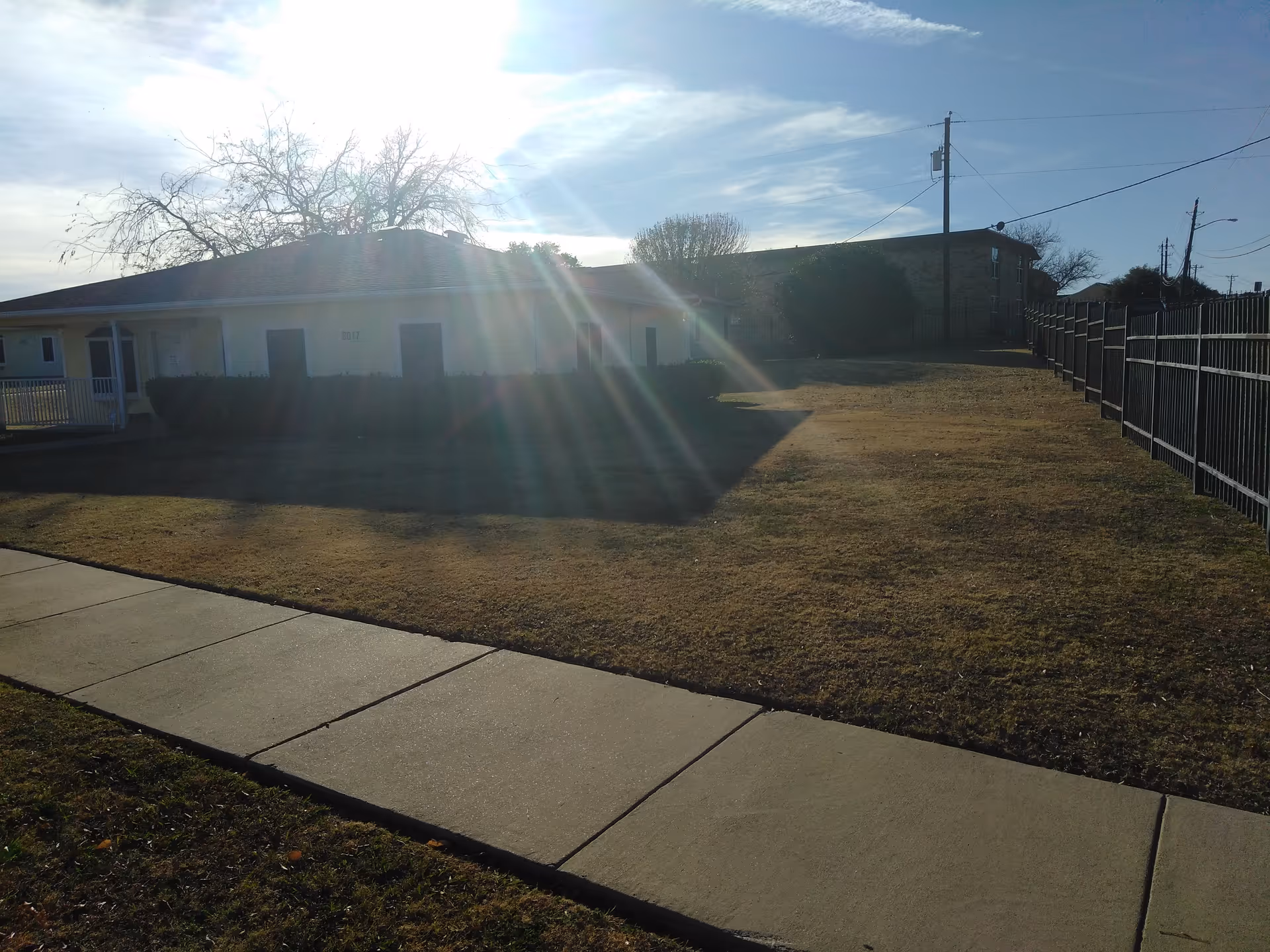 Sunlit single-story Tremont Terrace exterior with sidewalk, lawn, and a tall fence.