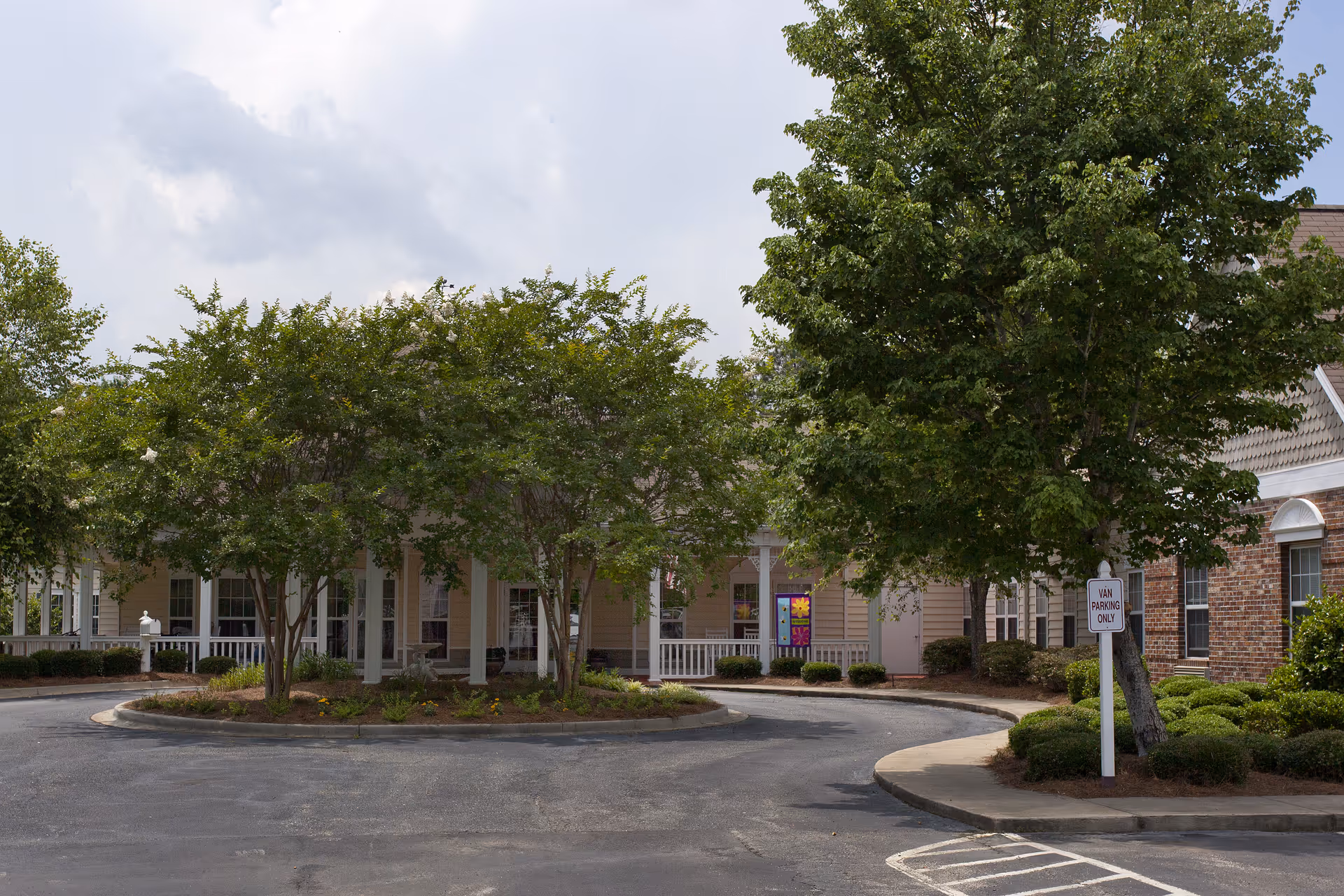 Exterior view of Oaks at Maple Ridge facility showing a circular driveway with landscaped island featuring several trees and bushes. The building has a covered porch with white railings and a mix of brick and siding exterior walls. A sign indicating 'Van Parking Only' is visible near the driveway.