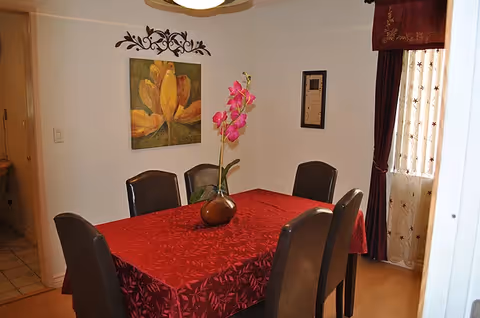 Dining room with a rectangular table draped in a red tablecloth, six chairs, a floral centerpiece, wall art, and curtained window.
