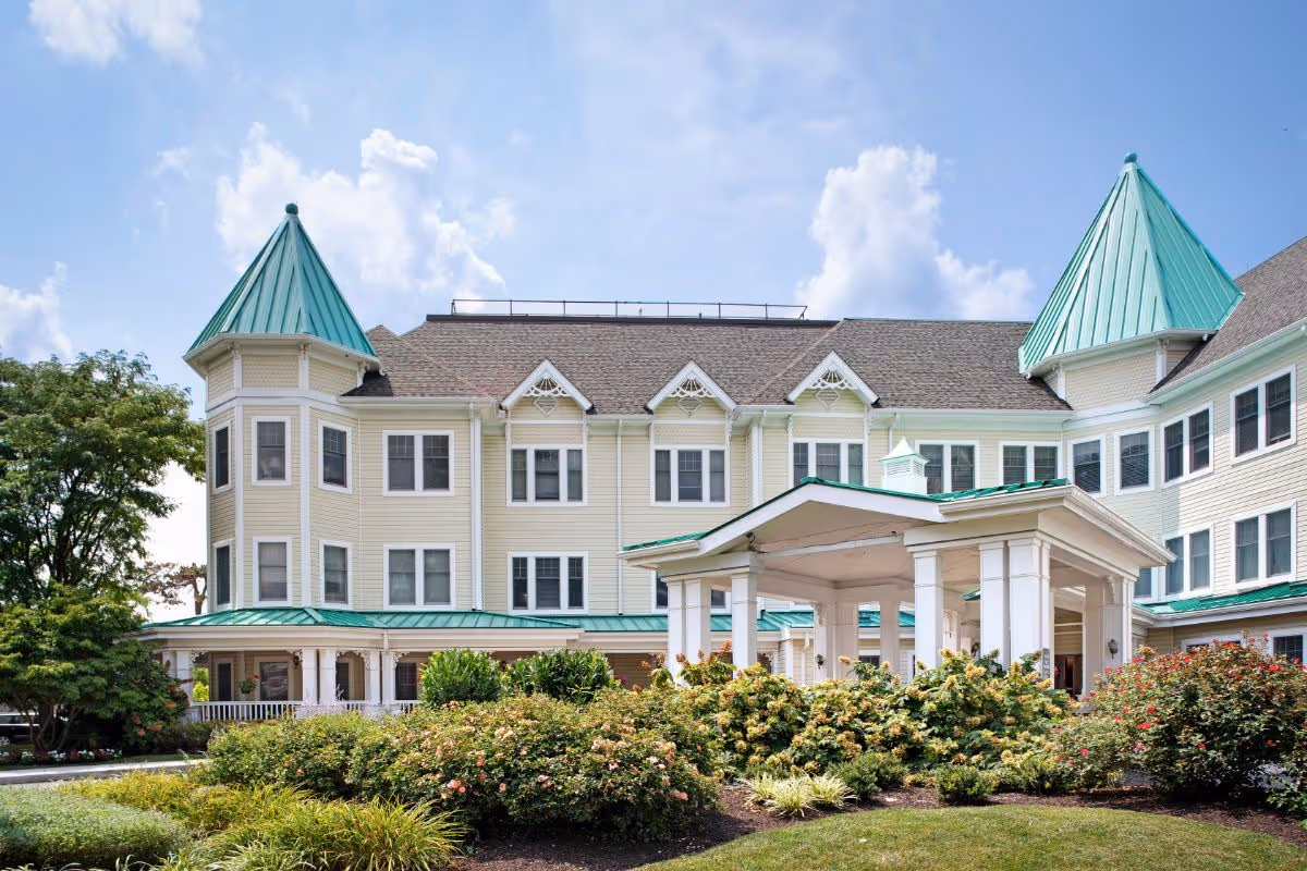 Exterior view of a large senior living facility building with beige siding and green metal roofs on turrets and porch. The building is surrounded by well-maintained bushes and greenery under a partly cloudy blue sky.