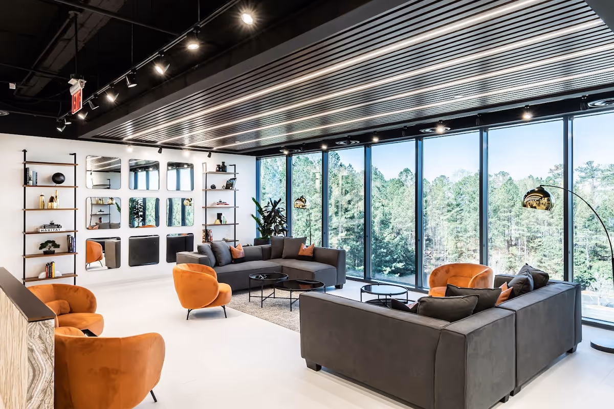 Modern senior living lounge with gray sofas, orange chairs, and windows overlooking trees.