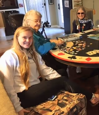 Three people sitting around a round table playing a jigsaw puzzle in a cozy room. The table has a playing card suit design on its edge. One young woman with long blonde hair is sitting on a couch smiling at the camera, while two older women are seated at the table working on the puzzle.