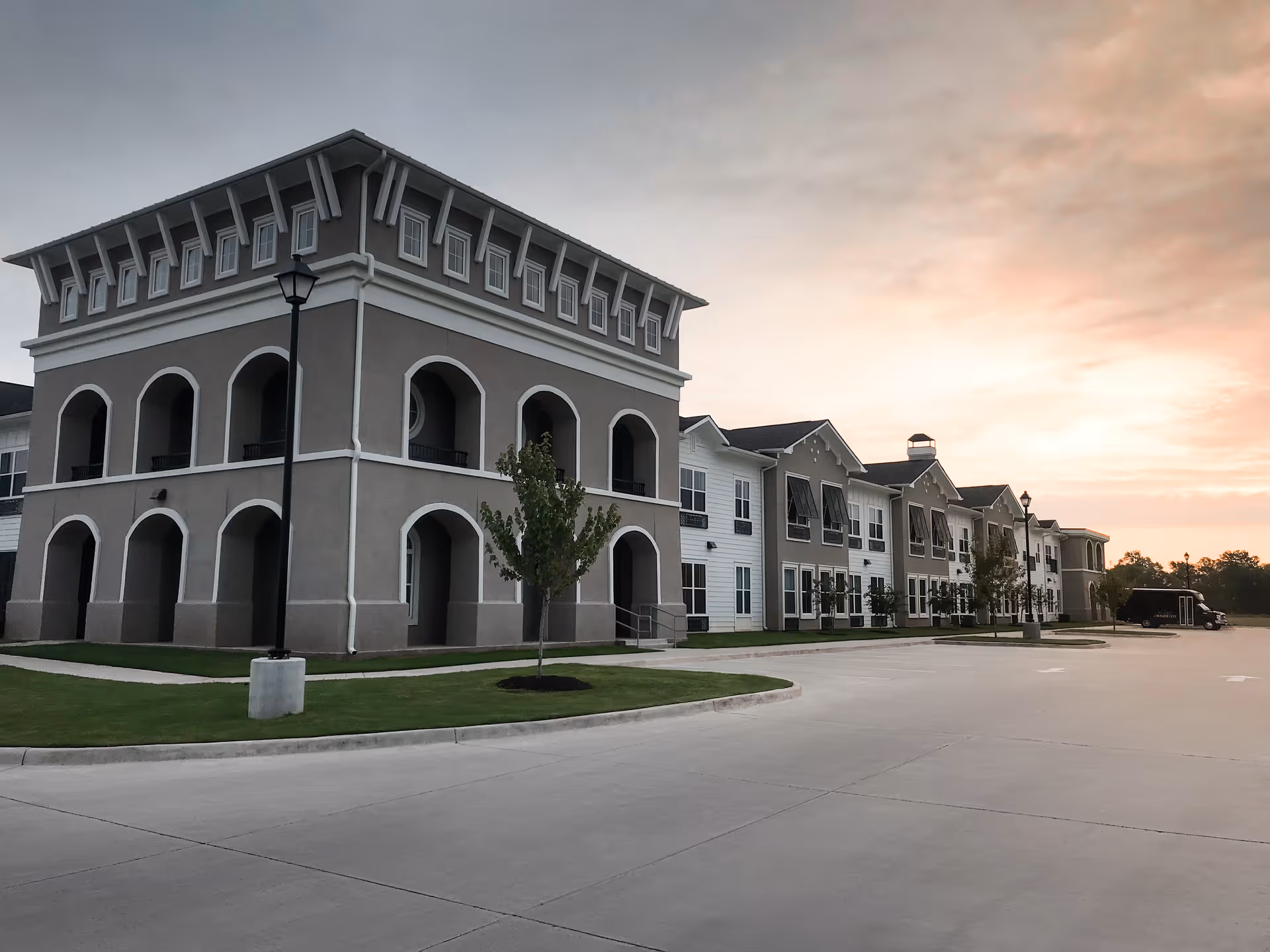 Front exterior of a multi-story senior living building featuring arched balconies and a parking area at sunset.