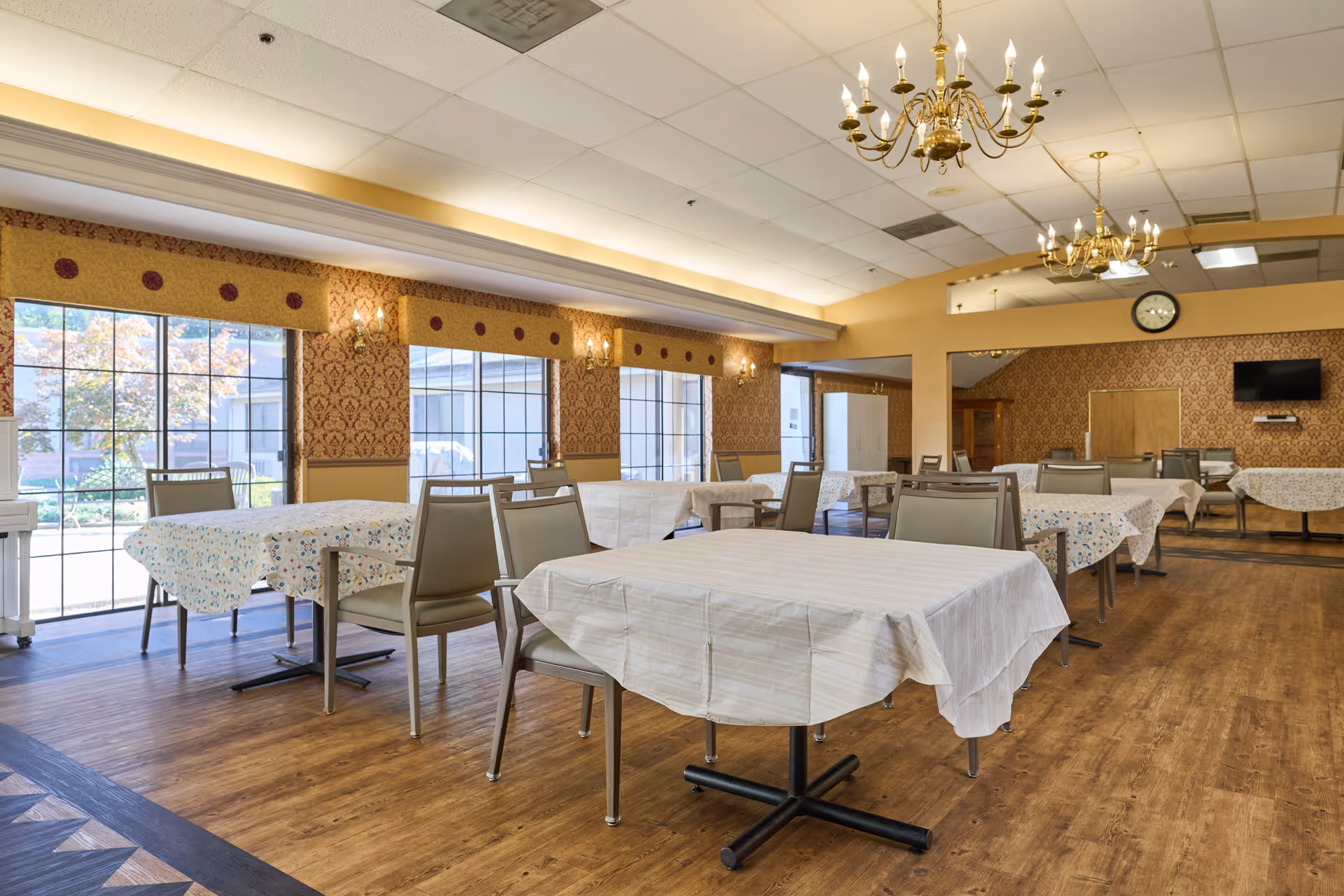 Spacious dining room with covered tables, chandeliers, and large windows in a senior living facility.