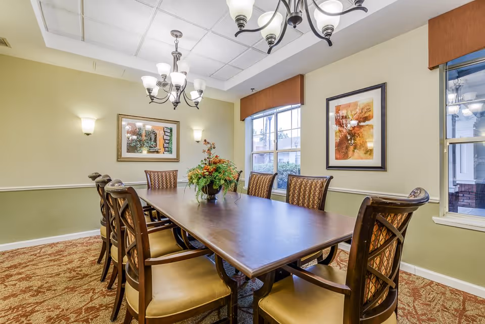 A well-lit dining room with a long wooden table surrounded by eight cushioned chairs. The room features two large windows with brown valances, two framed paintings on the walls, a floral centerpiece on the table, and a chandelier hanging from the ceiling. The walls are painted in a two-tone style with a light green lower half and beige upper half.