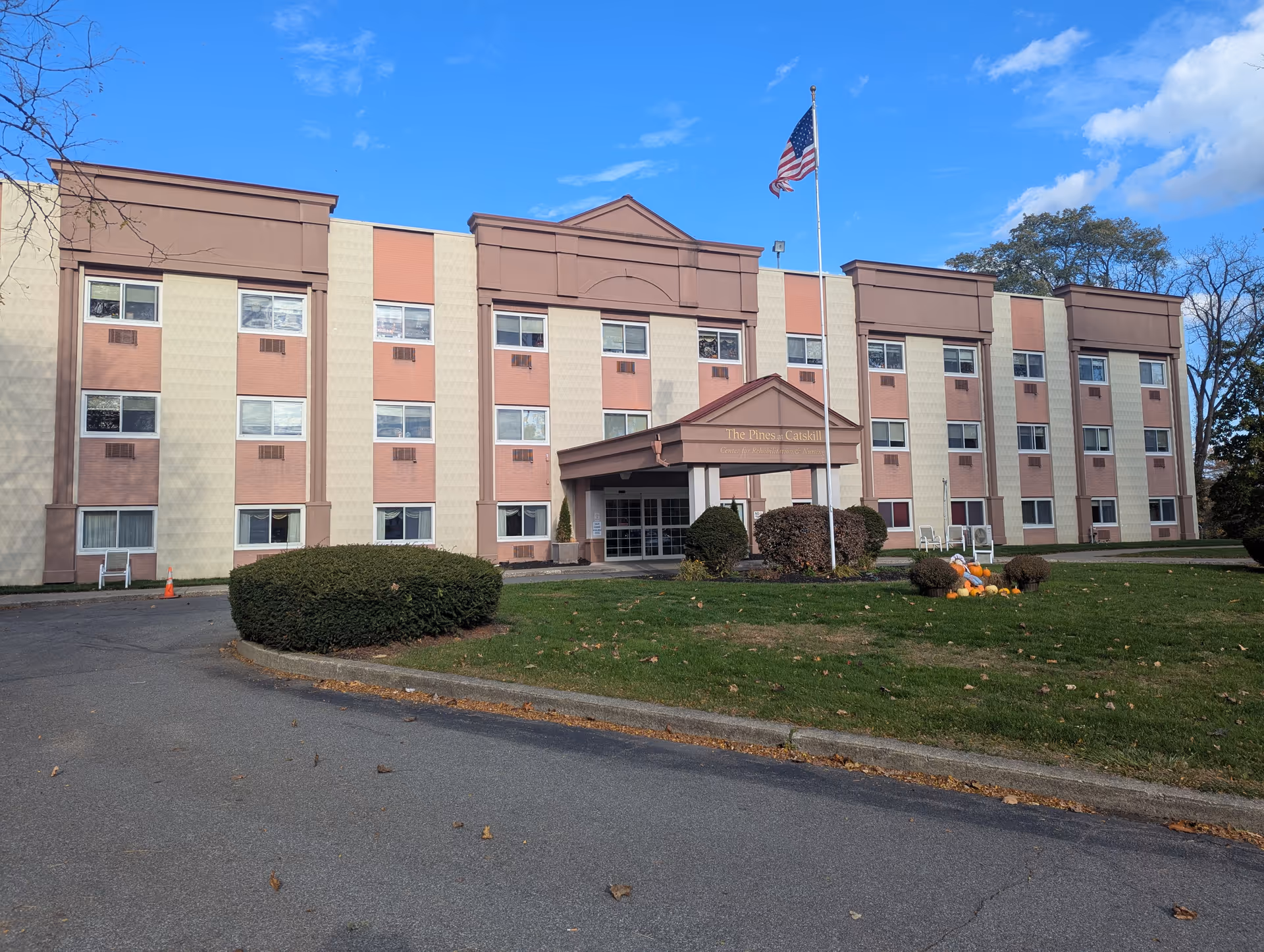Multi-story senior living building with beige-pink exterior, entrance canopy, flagpole, and landscaped lawn.