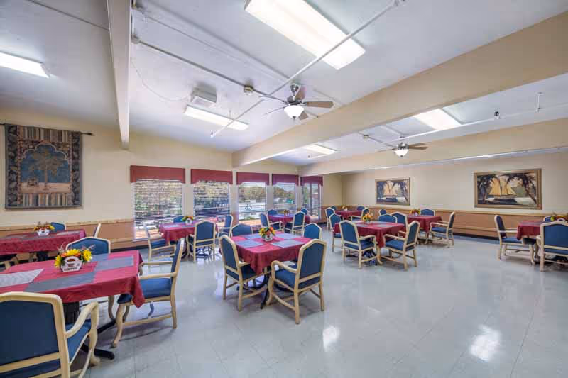Bright dining hall with red tablecloths, blue chairs, and ceiling fans in a senior facility.