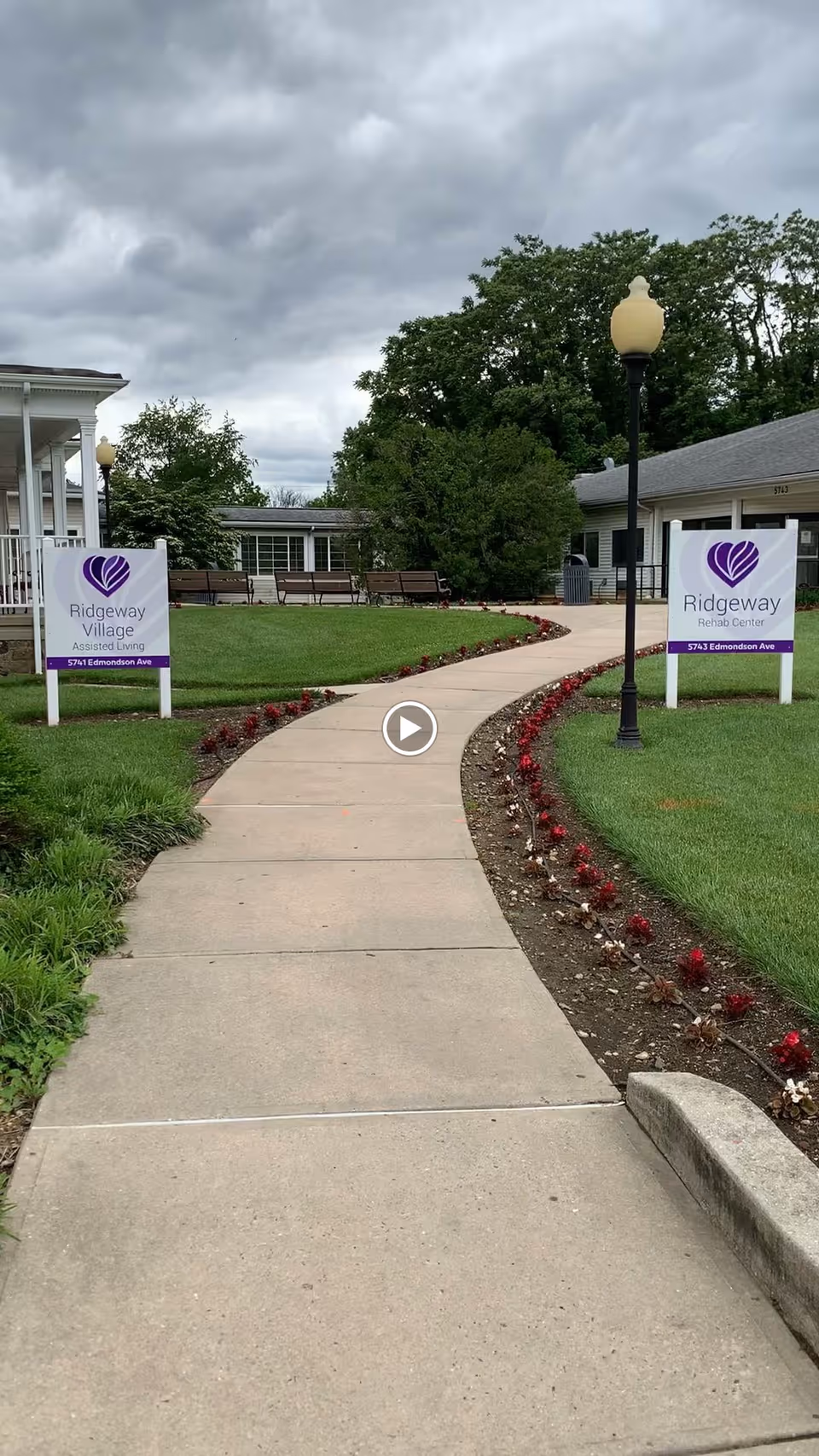 Curved concrete walkway through landscaped lawn and flower beds leading to the entrance of a senior living building with Ridgeway signs and lamp posts under a cloudy sky.