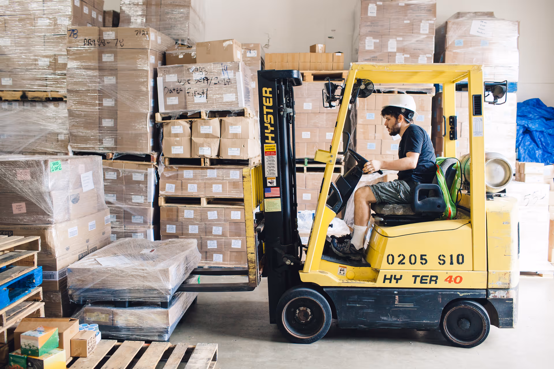 Worker operates a yellow forklift among stacked pallets in an indoor warehouse.