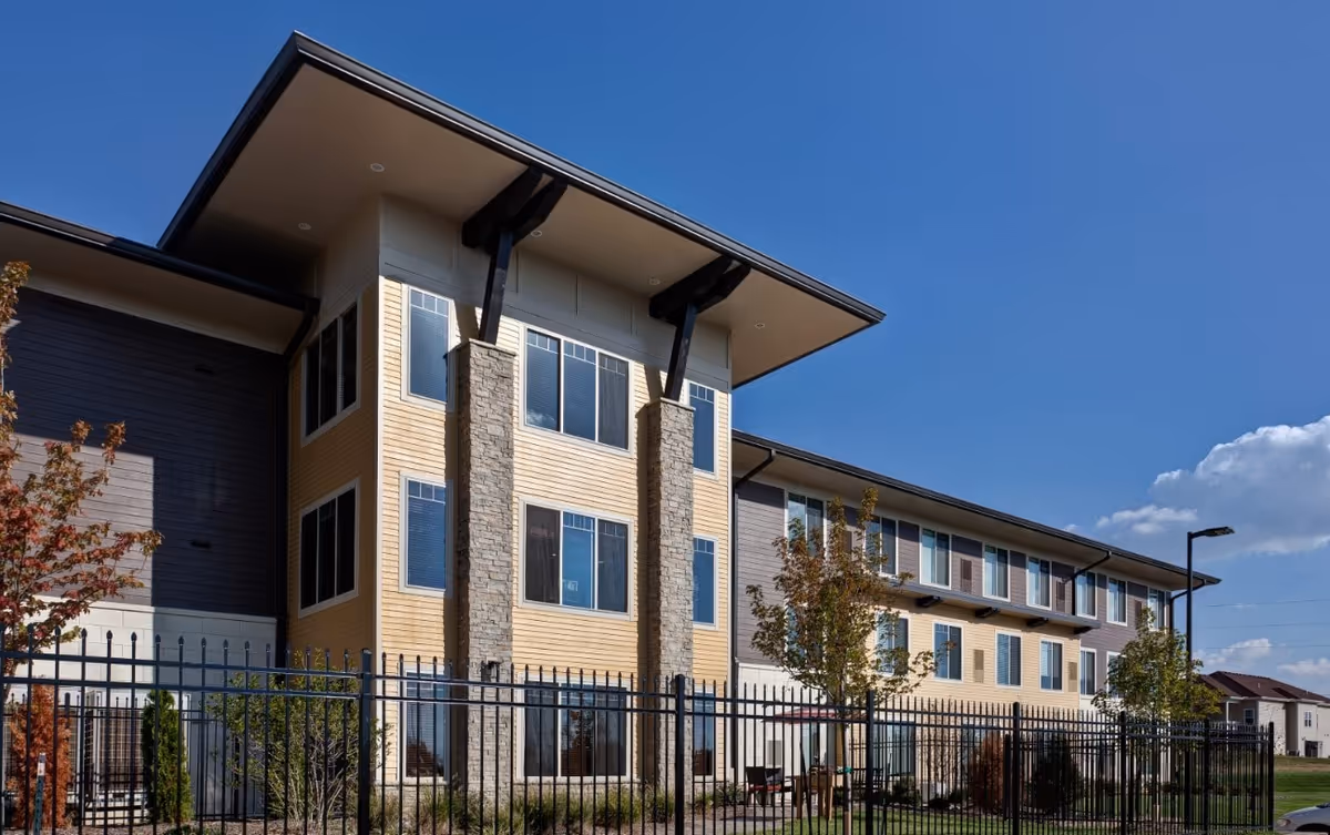 Exterior view of a modern assisted living building with beige siding, stone columns, and a fence.