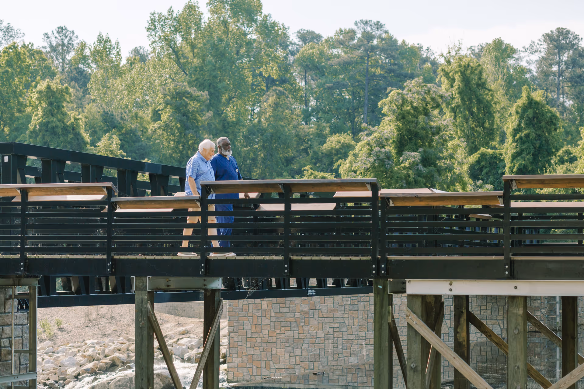 Two elderly men stand on a wooden bridge overlooking a river and trees.