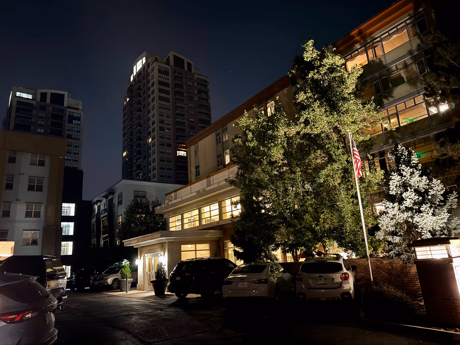 Night exterior of a multi-story senior living building with a lit entrance and parked cars.