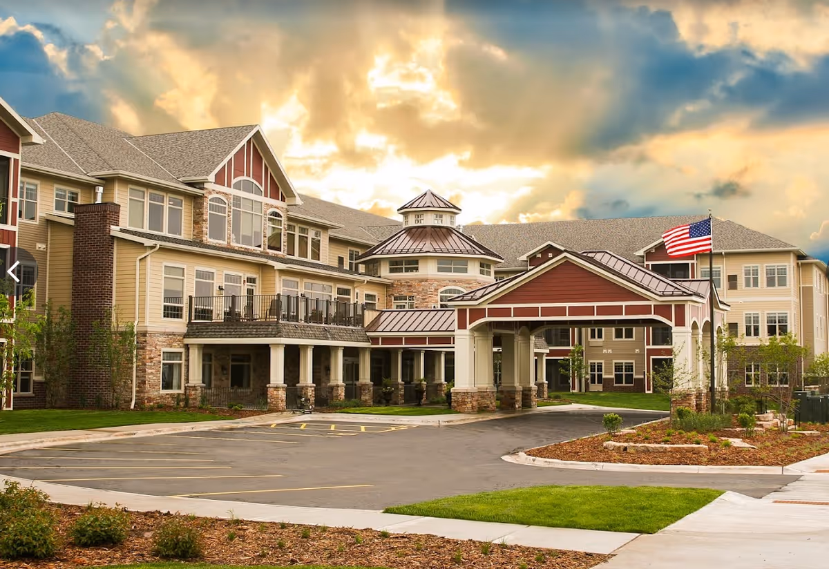 Exterior view of Parkside Post Acute and Rehabilitation Center, a large multi-story building with beige and red siding, stone accents, and a covered entrance. The American flag is flying on a flagpole near the entrance, and the sky is partly cloudy with a warm glow from the setting or rising sun.