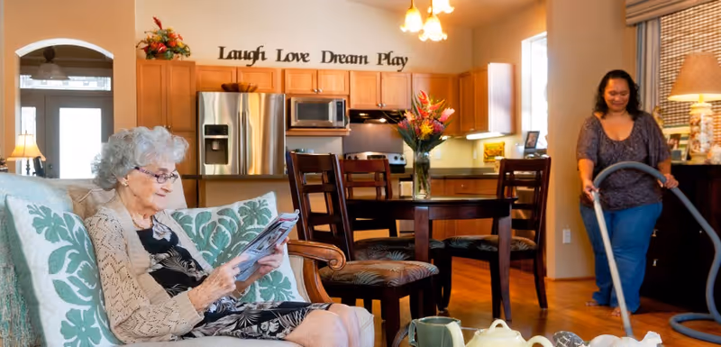 An elderly woman sits on a sofa reading a magazine while a staff member vacuums.