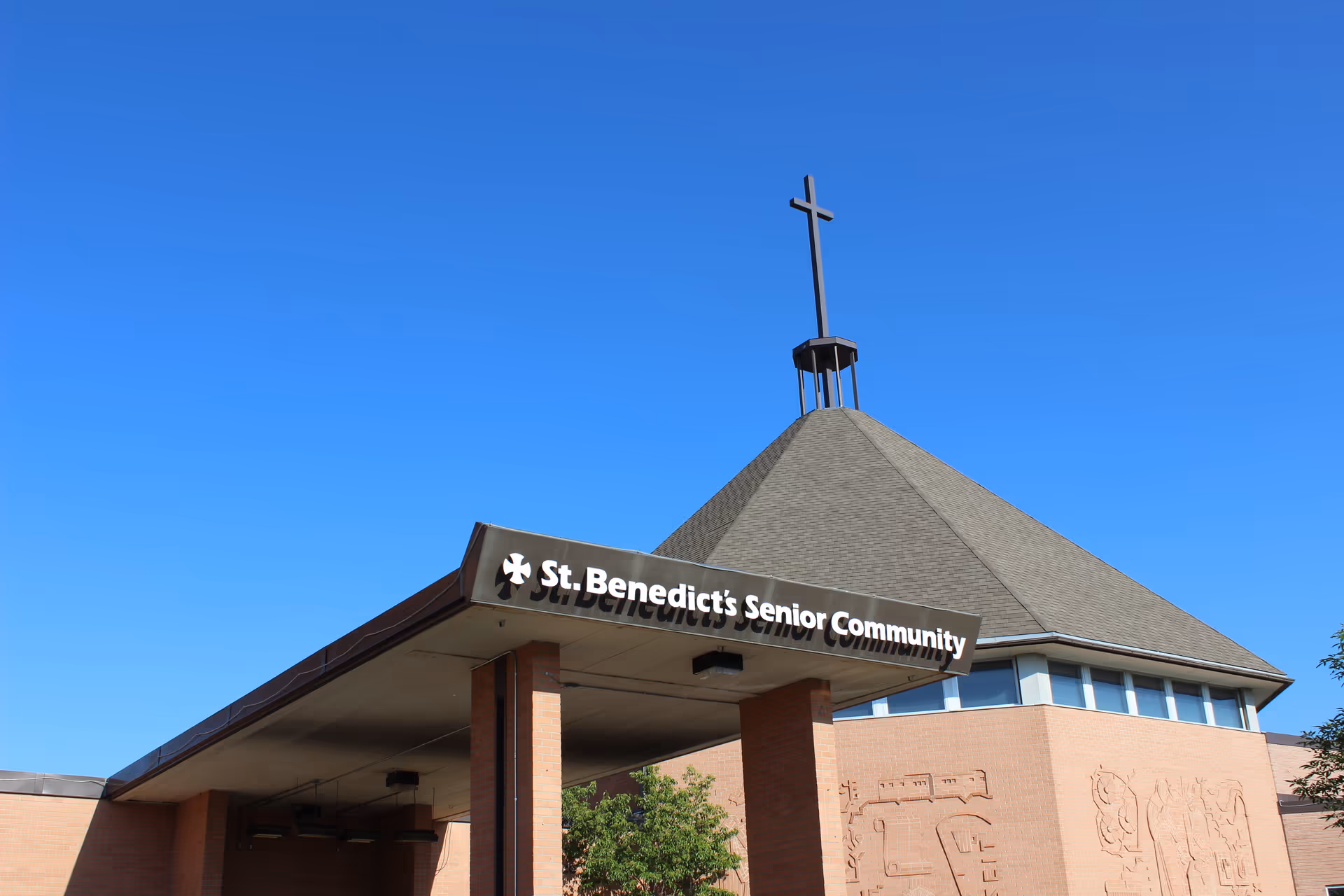 Exterior view of St. Benedict's Senior Community building featuring a brick structure with a large roof topped by a cross, under a clear blue sky.