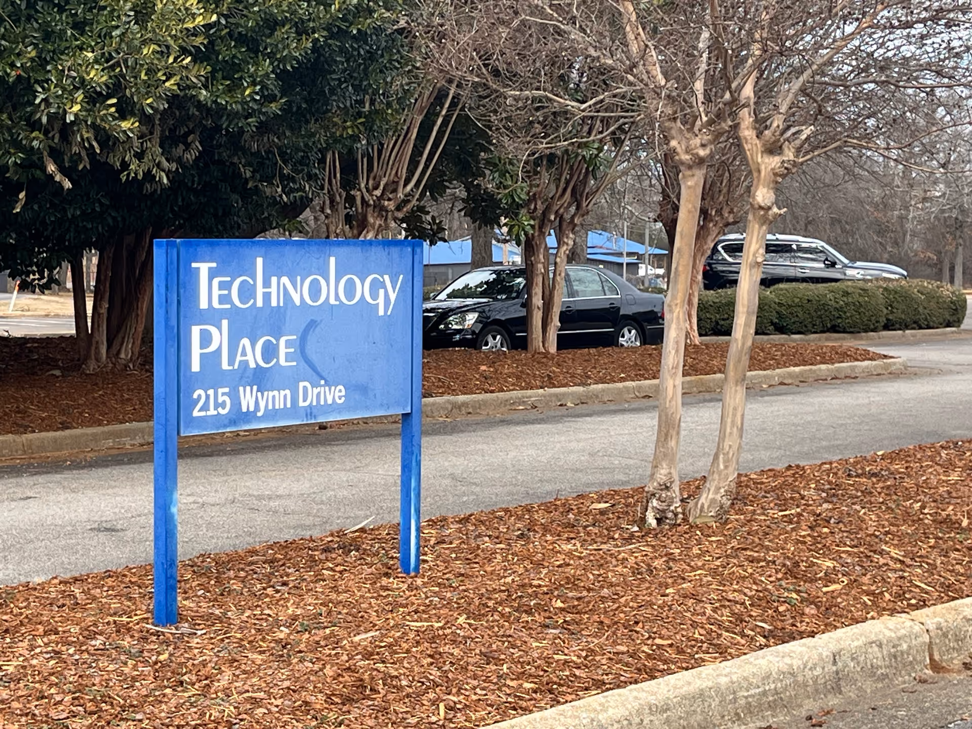 Blue Technology Place sign on mulch with trees and parked cars in the background.