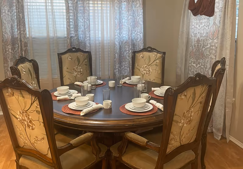 Round wooden dining table with place settings and ornate upholstered chairs in a curtained dining room.