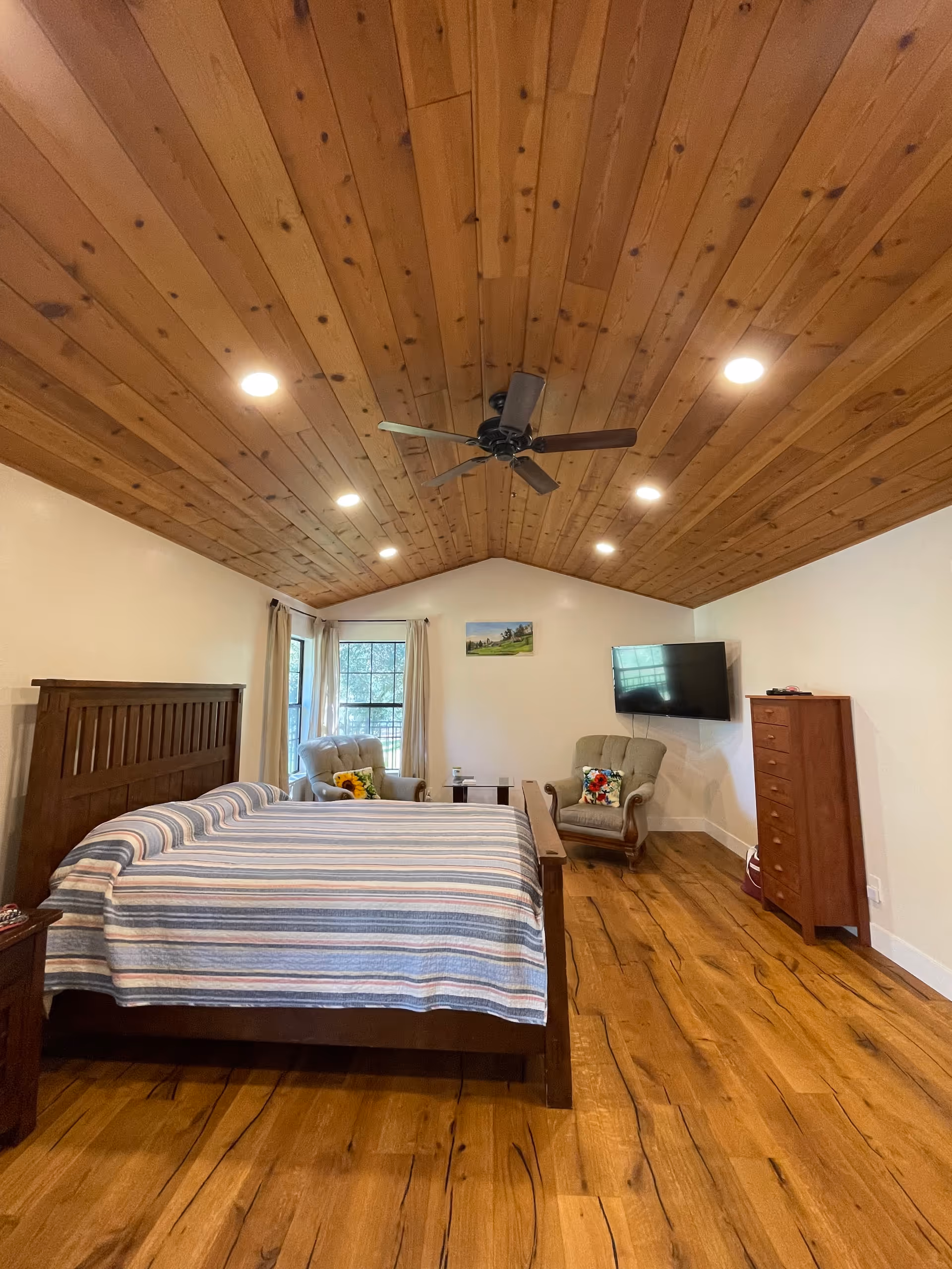 A spacious bedroom with a wooden vaulted ceiling, a large bed with a striped cover, two armchairs by a window, a wall-mounted TV and a tall wooden dresser.