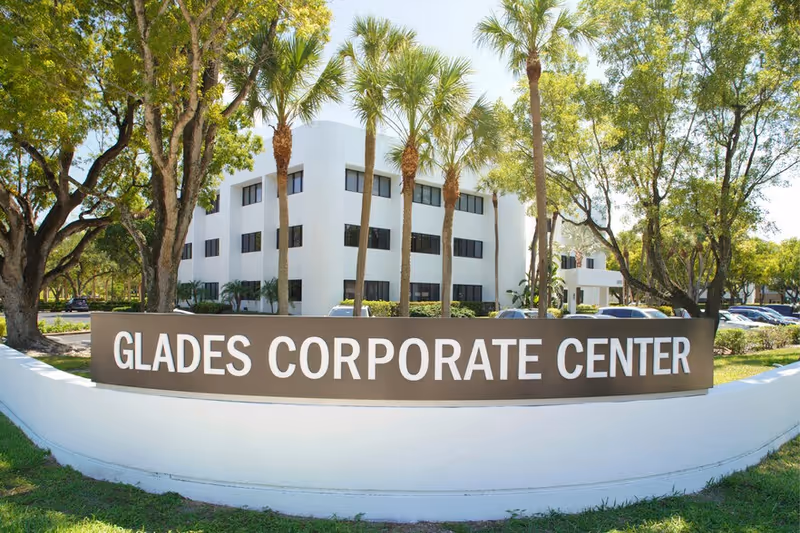 White multi-story building behind a curved sign reading GLADES CORPORATE CENTER, surrounded by palm trees.