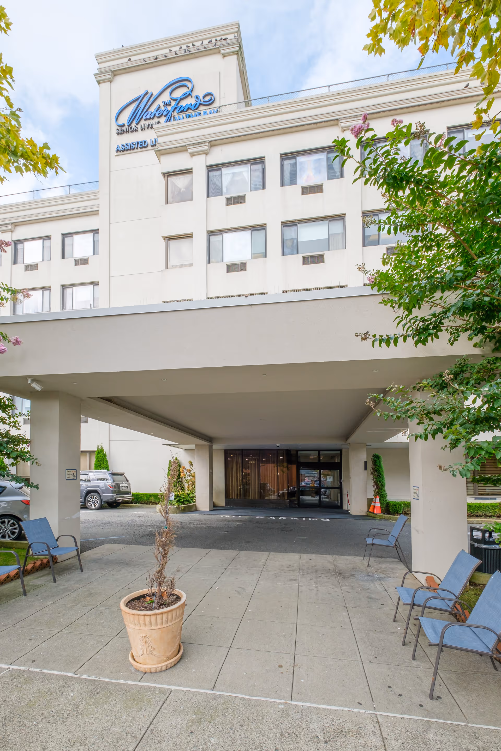 Exterior view of The Waterford on the Bay Senior Living building showing the entrance area with a covered drop-off zone, several blue chairs arranged on the sidewalk, a potted plant, and some parked cars nearby. The building has multiple windows and a sign with the facility name on the upper part.