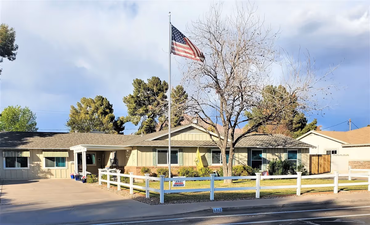 Exterior view of Hazelwood Manor Assisted Living, a single-story building with beige siding and a brown roof. There is a white fence surrounding a small grassy area with a leafless tree and an American flag on a flagpole. The driveway and sidewalk are visible in front of the building under a partly cloudy sky.