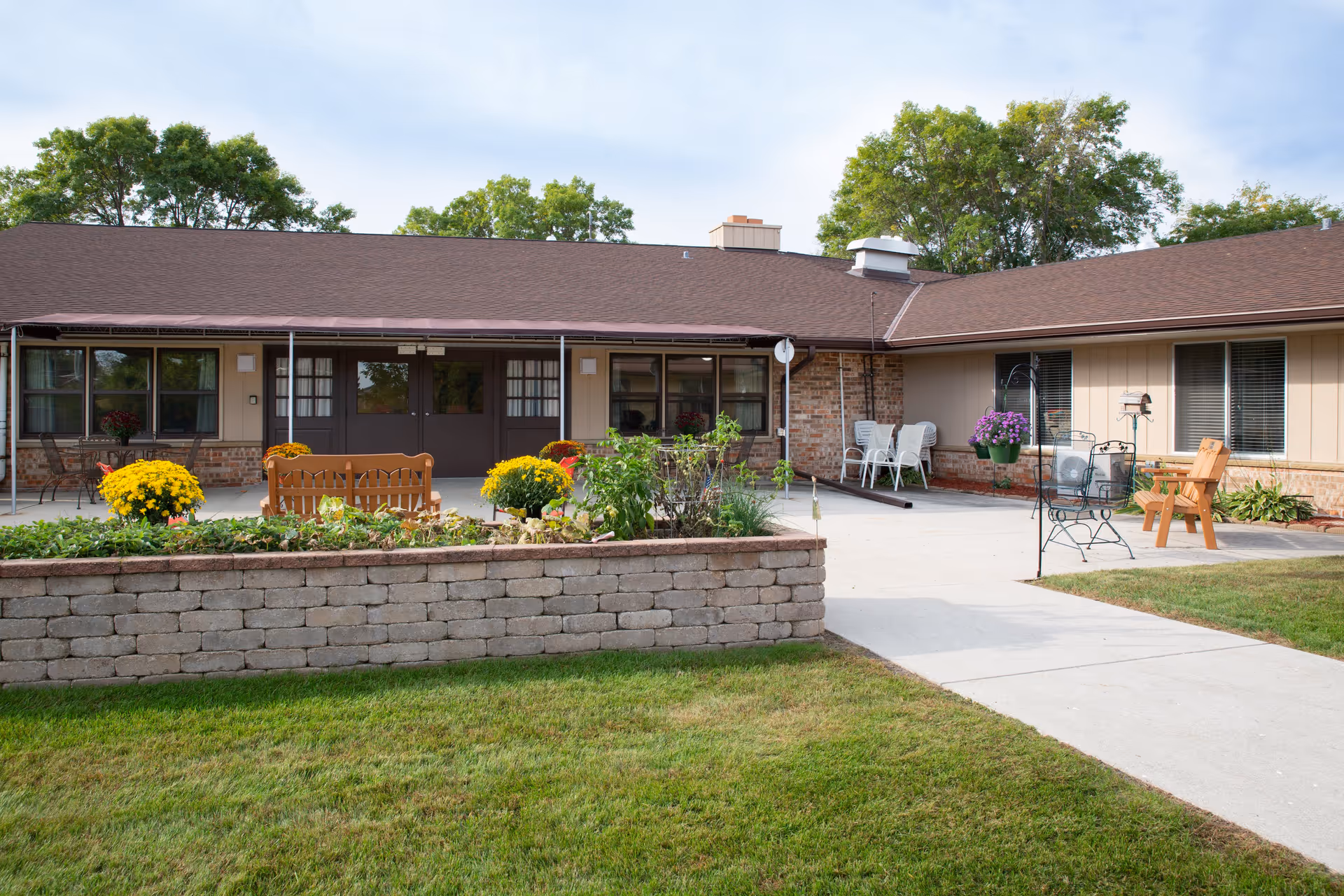 Exterior view of a single-story senior living facility building with a brown roof and beige walls. There is a raised garden bed with yellow flowers and greenery in front of the building. Several chairs and benches are placed on the concrete patio area. Trees are visible in the background under a partly cloudy sky.