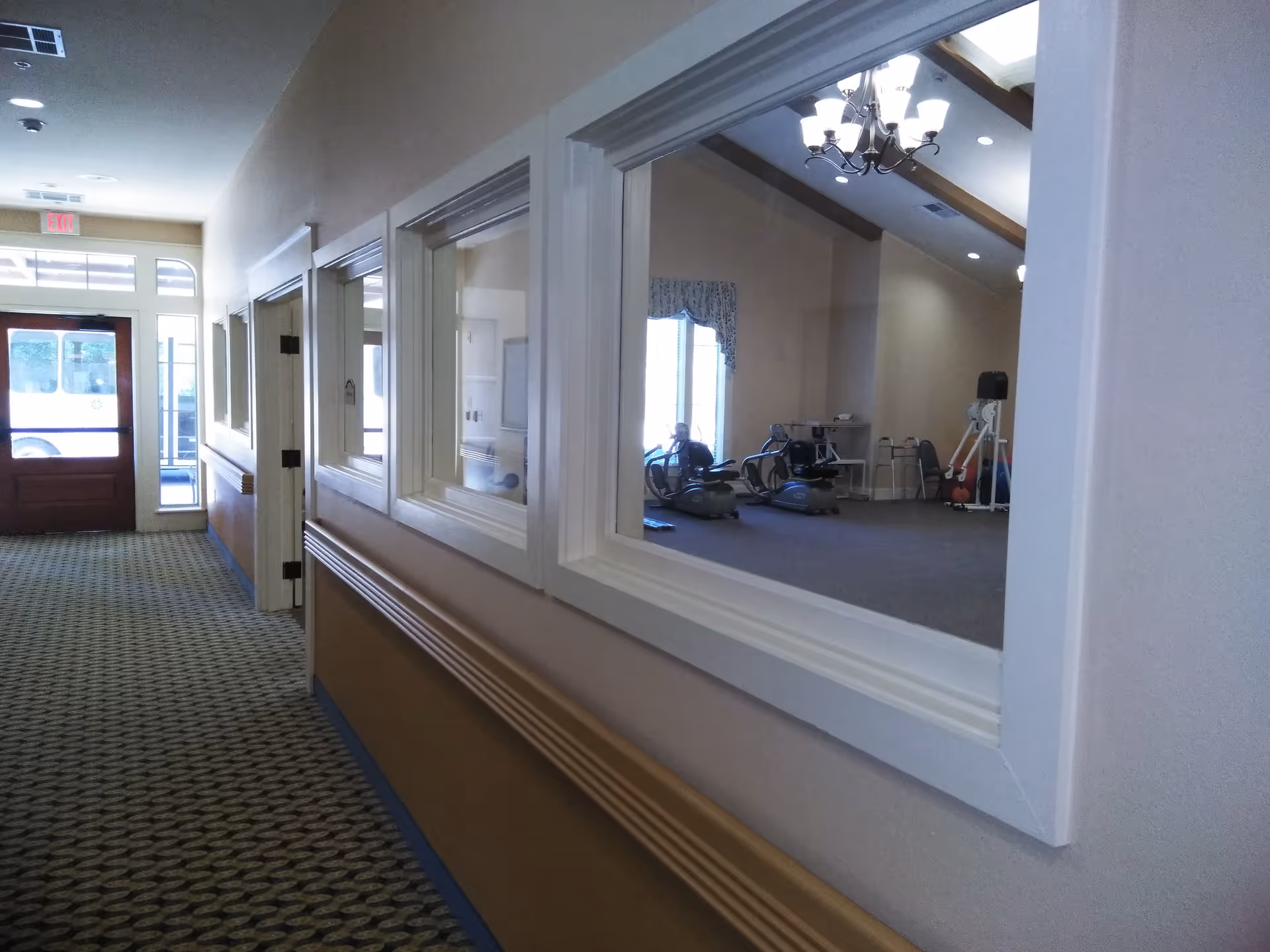 Interior hallway of a senior living facility with exercise room visible through windows.