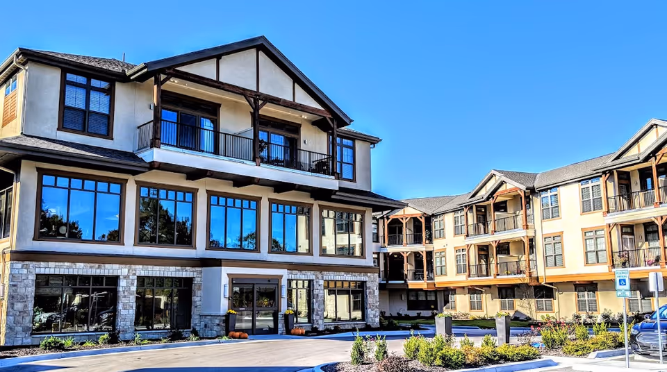Front exterior of a multi-story senior living building with large windows, balconies, and a landscaped entrance under a clear blue sky.