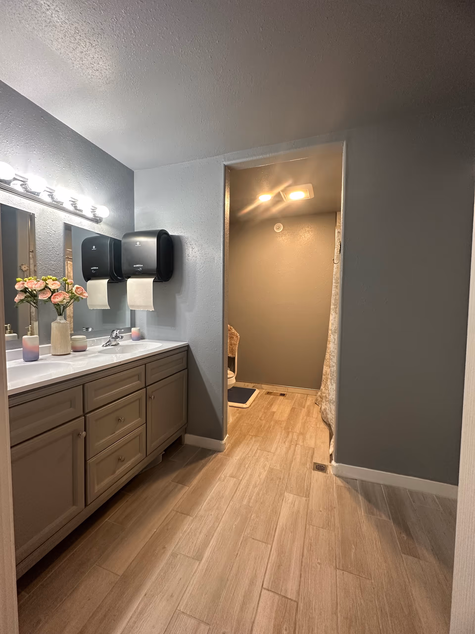 Bathroom with double sink vanity, mirrors, paper towel dispensers, and a doorway to a separate room.