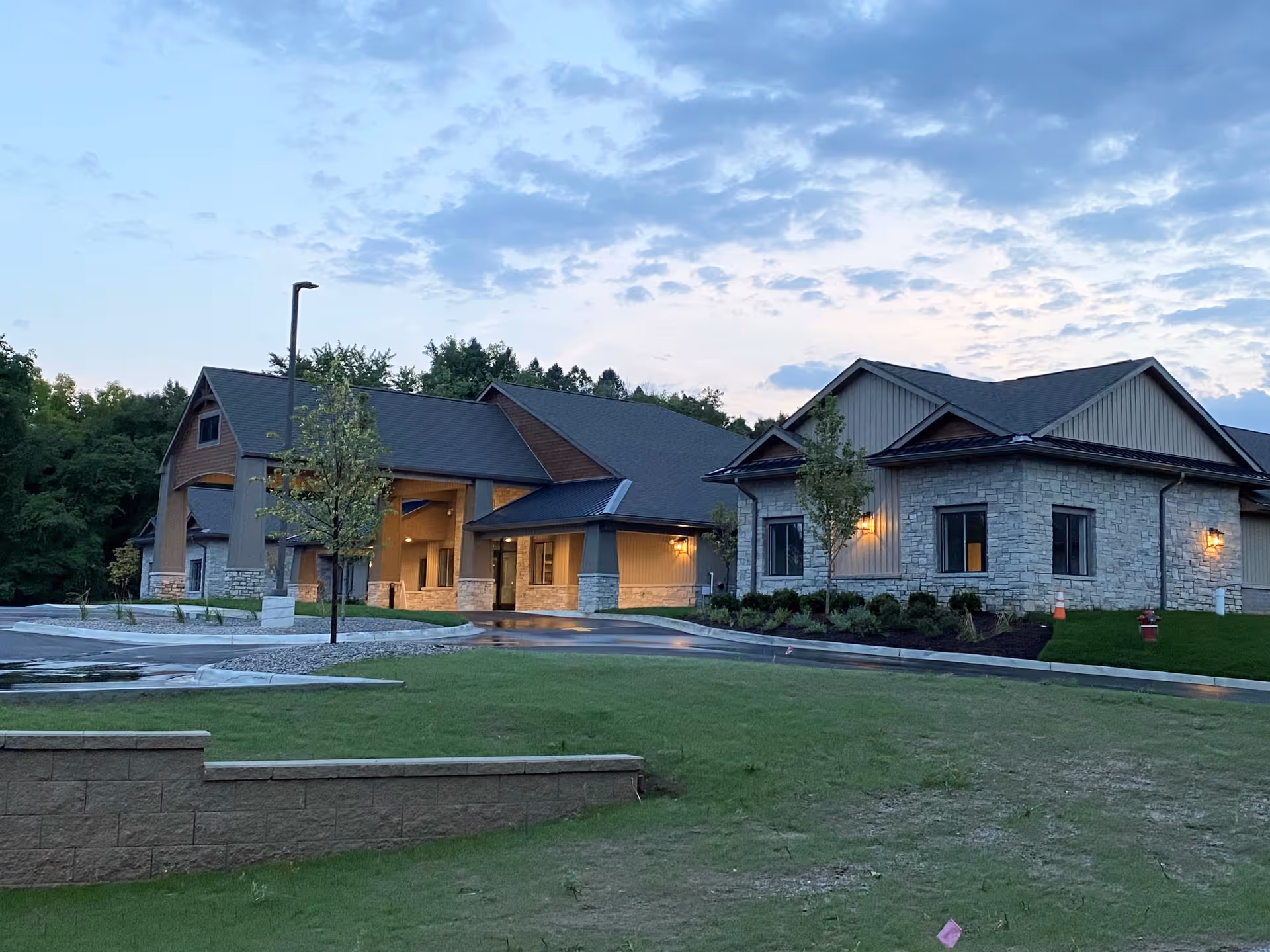 Front exterior of a single-story senior living facility with a stone facade, lit entrance canopy, and landscaped lawn at dusk.