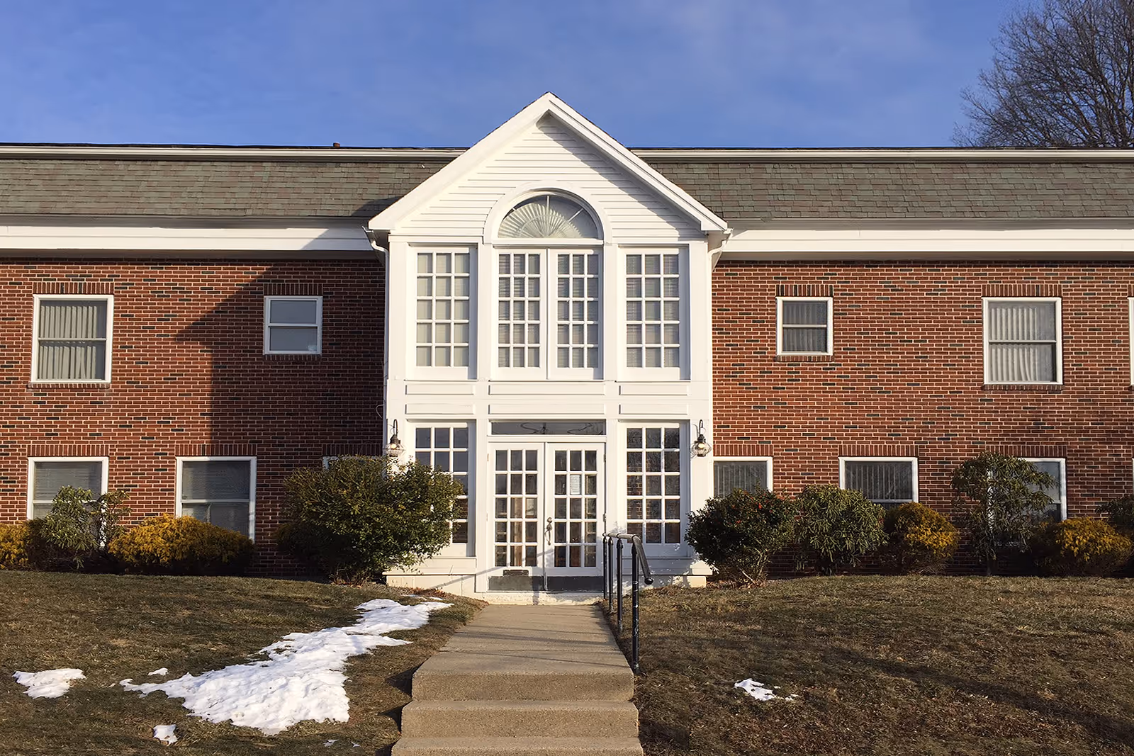 Brick building with a white central entrance and steps, shrubs, and patches of snow.