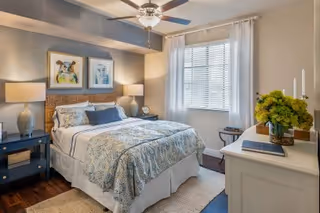 A cozy bedroom with a neatly made bed featuring patterned bedding and multiple pillows. The room has a ceiling fan with a light, two bedside tables with lamps, framed artwork above the bed, a window with white curtains, and a white dresser with a vase of green flowers and books on top.
