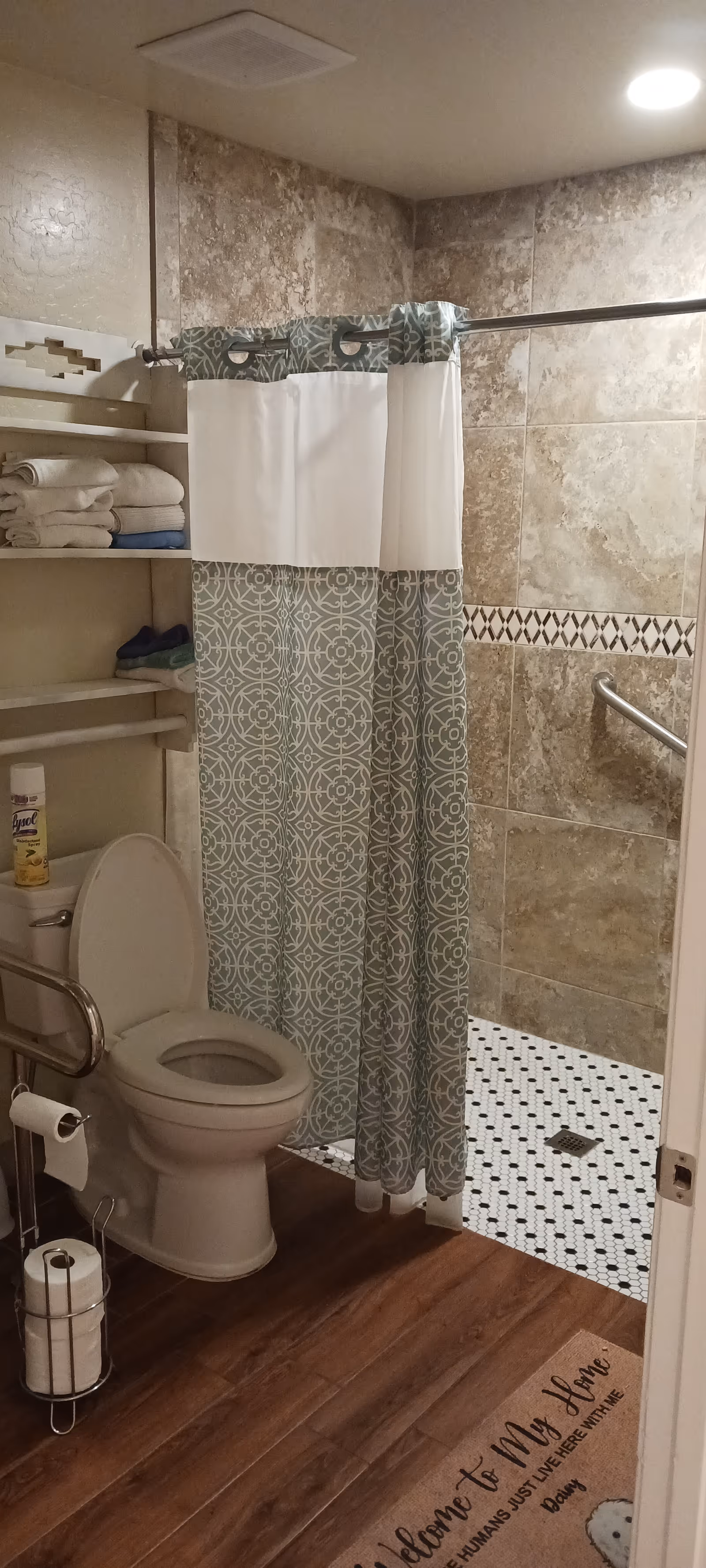 Senior living bathroom with shower curtain, toilet, towels on shelves, grab bars, wood-like flooring.