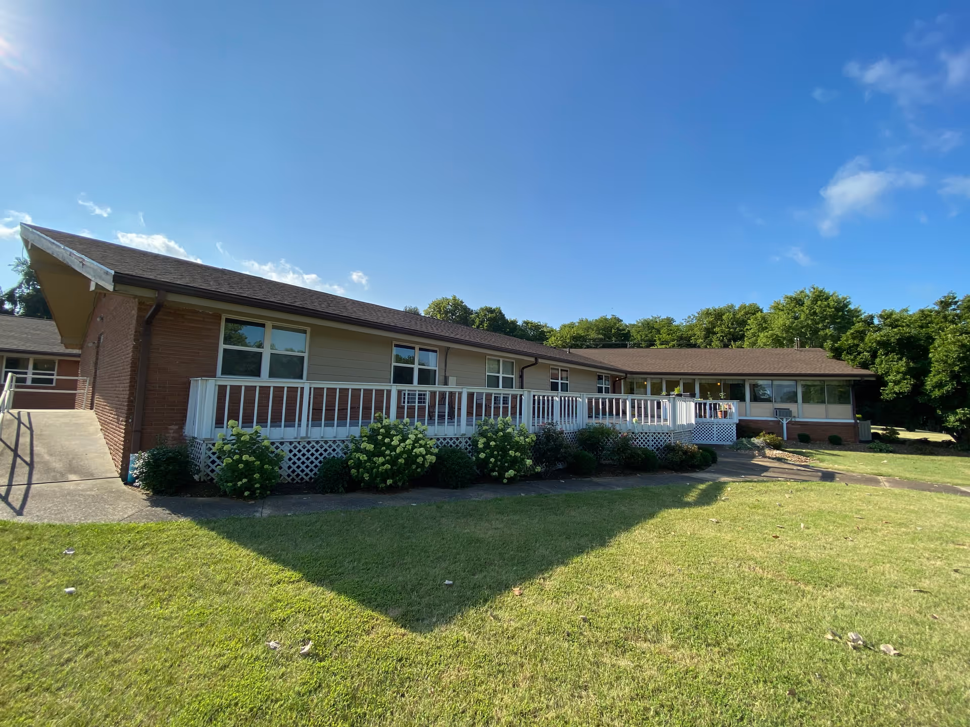 Long, single-story senior living facility exterior with white railings, brick walls, and a lush lawn.