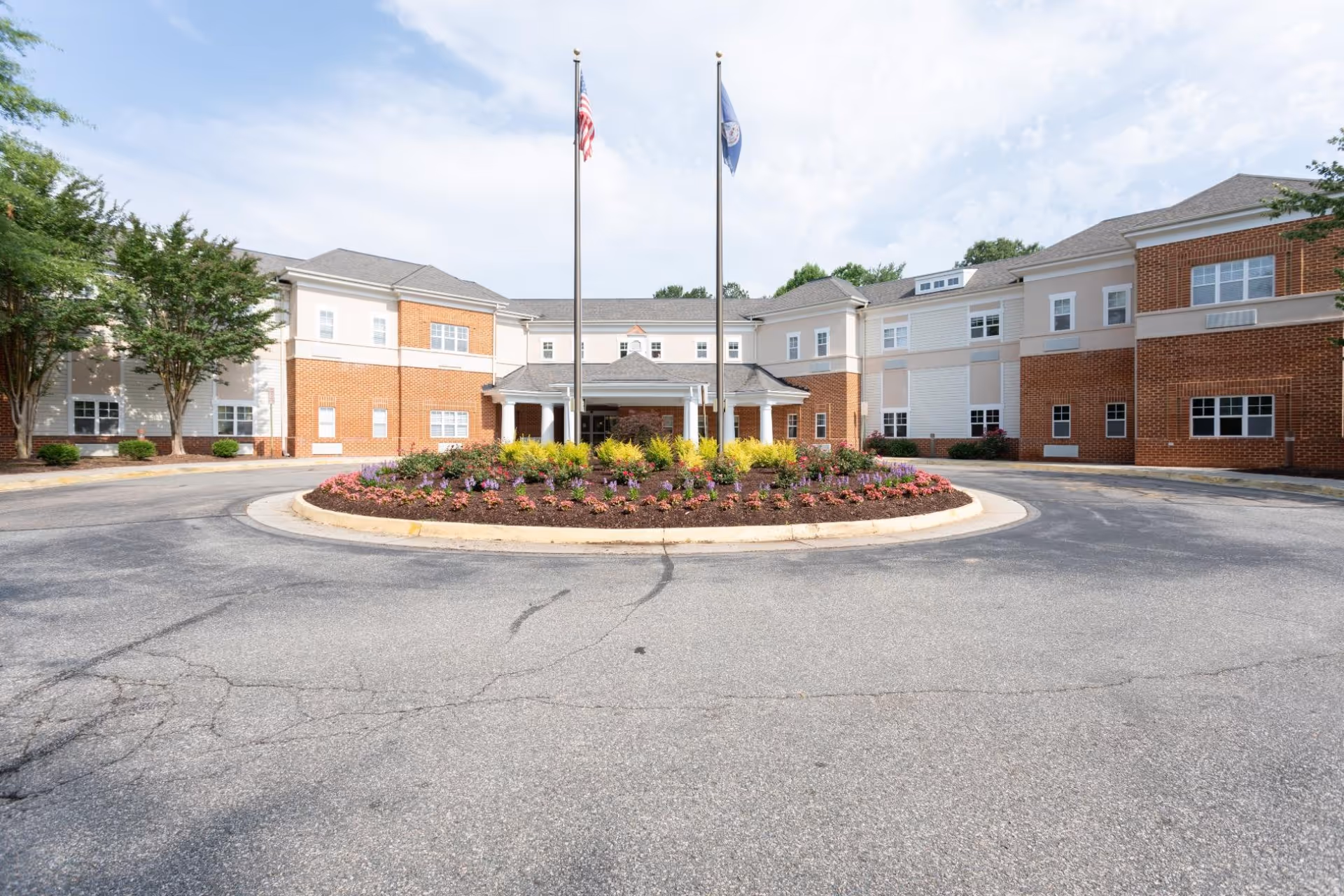 Front exterior view of a two-story senior living facility building with a circular driveway and a landscaped roundabout featuring colorful flowers and shrubs. Two flagpoles with the American flag and another flag stand in front of the entrance under a covered porch. The building has a combination of brick and light-colored siding with multiple windows.