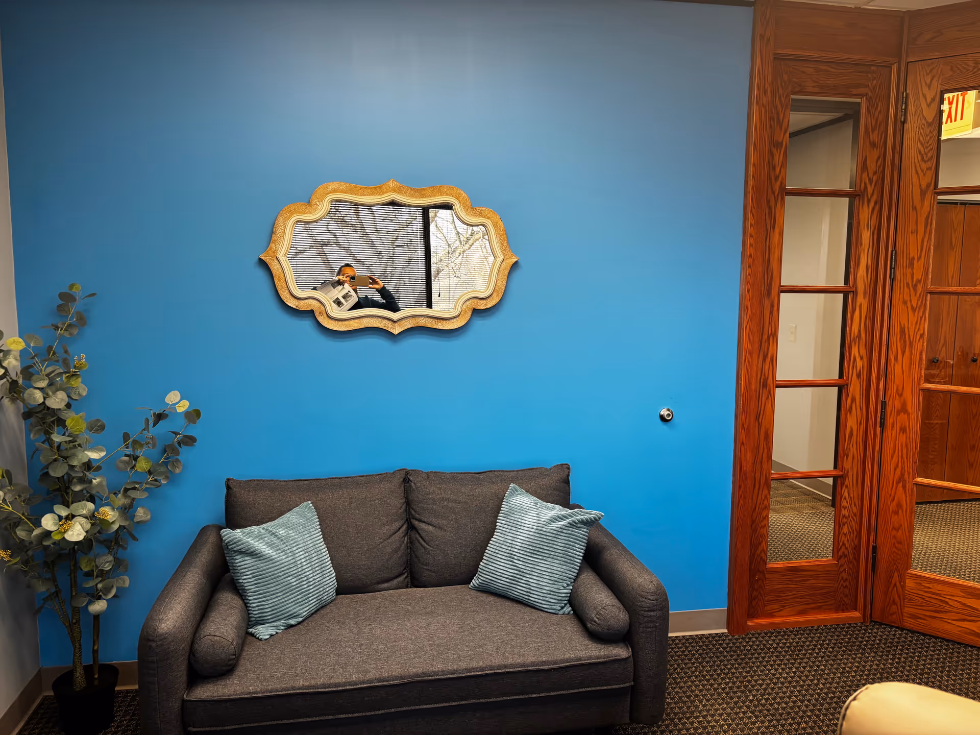 Blue-walled lounge with gray sofa, potted plant, decorative mirror, and wooden doors.
