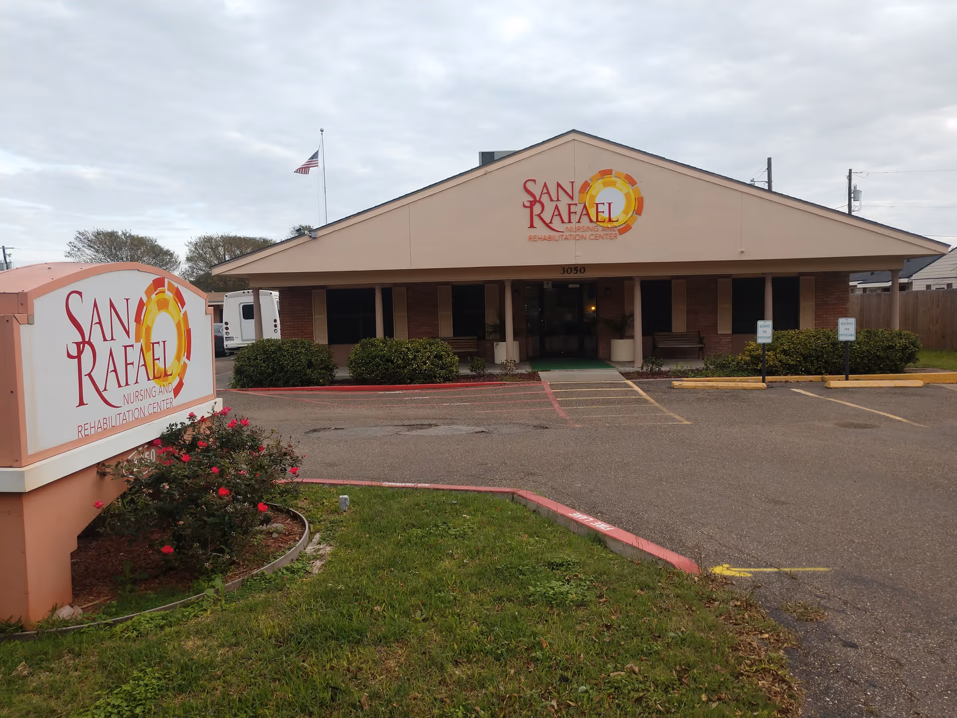 Front exterior view of San Rafael Nursing and Rehabilitation Center building with a sign displaying the facility name, a small parking area, some bushes, and an American flag on a pole against a cloudy sky.