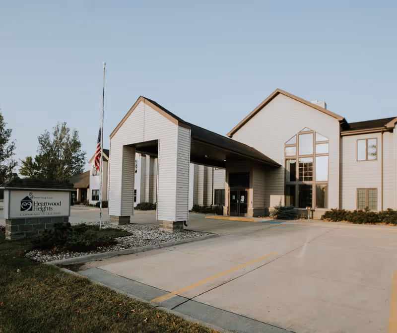 Exterior view of a senior living facility building with a covered entrance, large windows, and a sign that reads Heartwood Heights Senior Living Center. There is an American flag at half-mast near the entrance and a driveway leading up to the building.