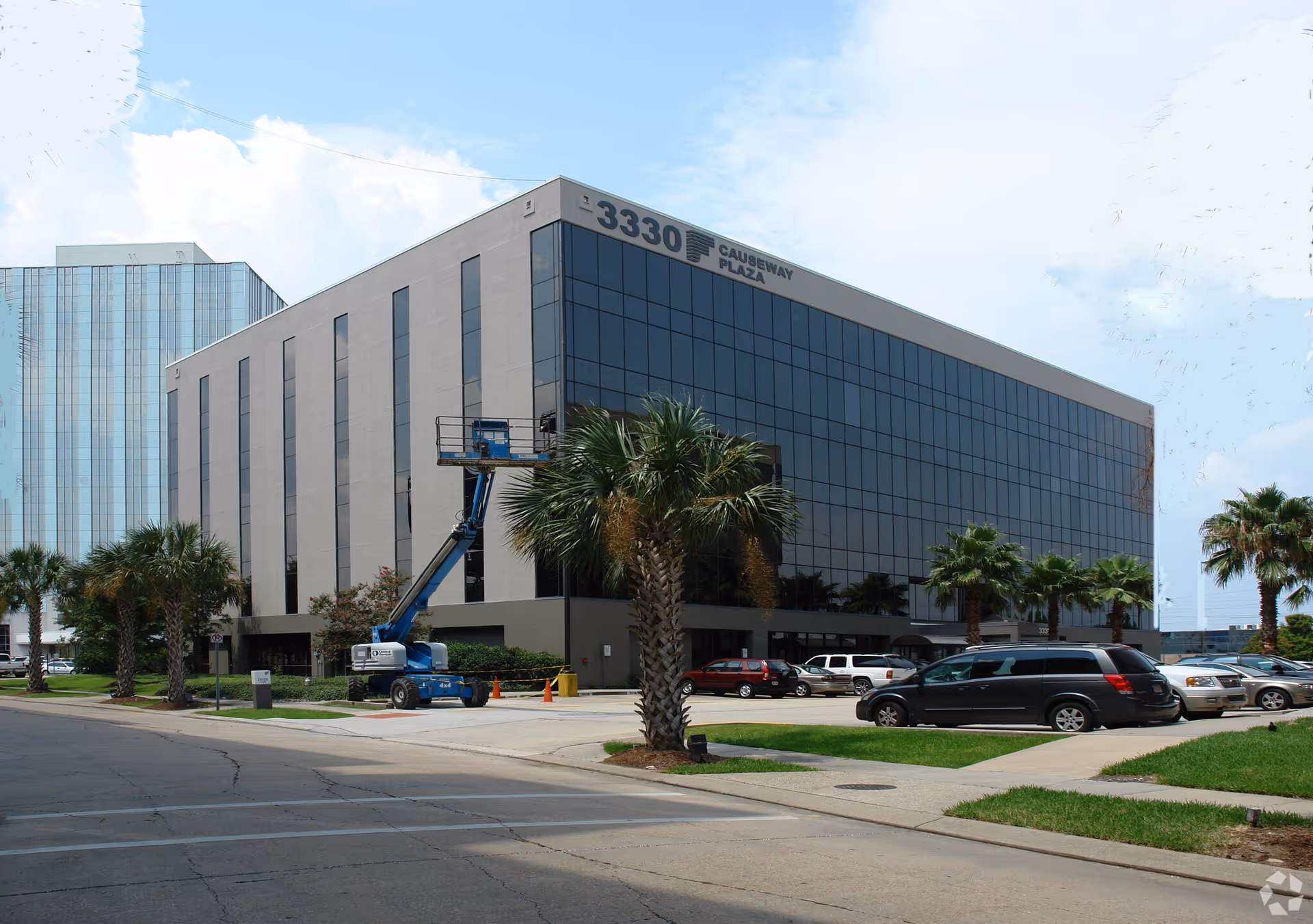 Exterior view of a modern glass-front building with palm trees, parked cars, and a construction lift.