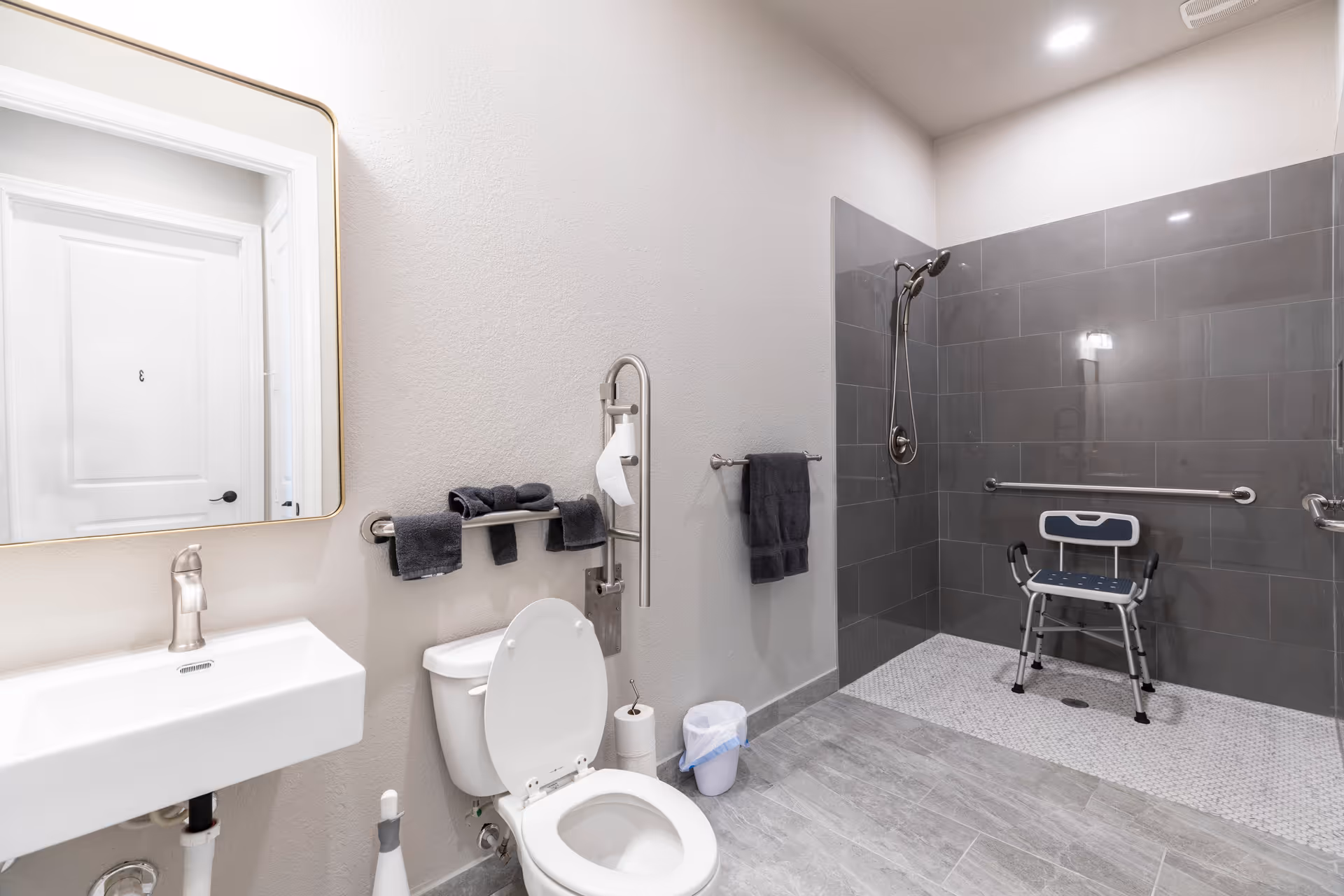Accessible bathroom with walk-in shower, grab bars, a toilet, sink, and a shower chair.