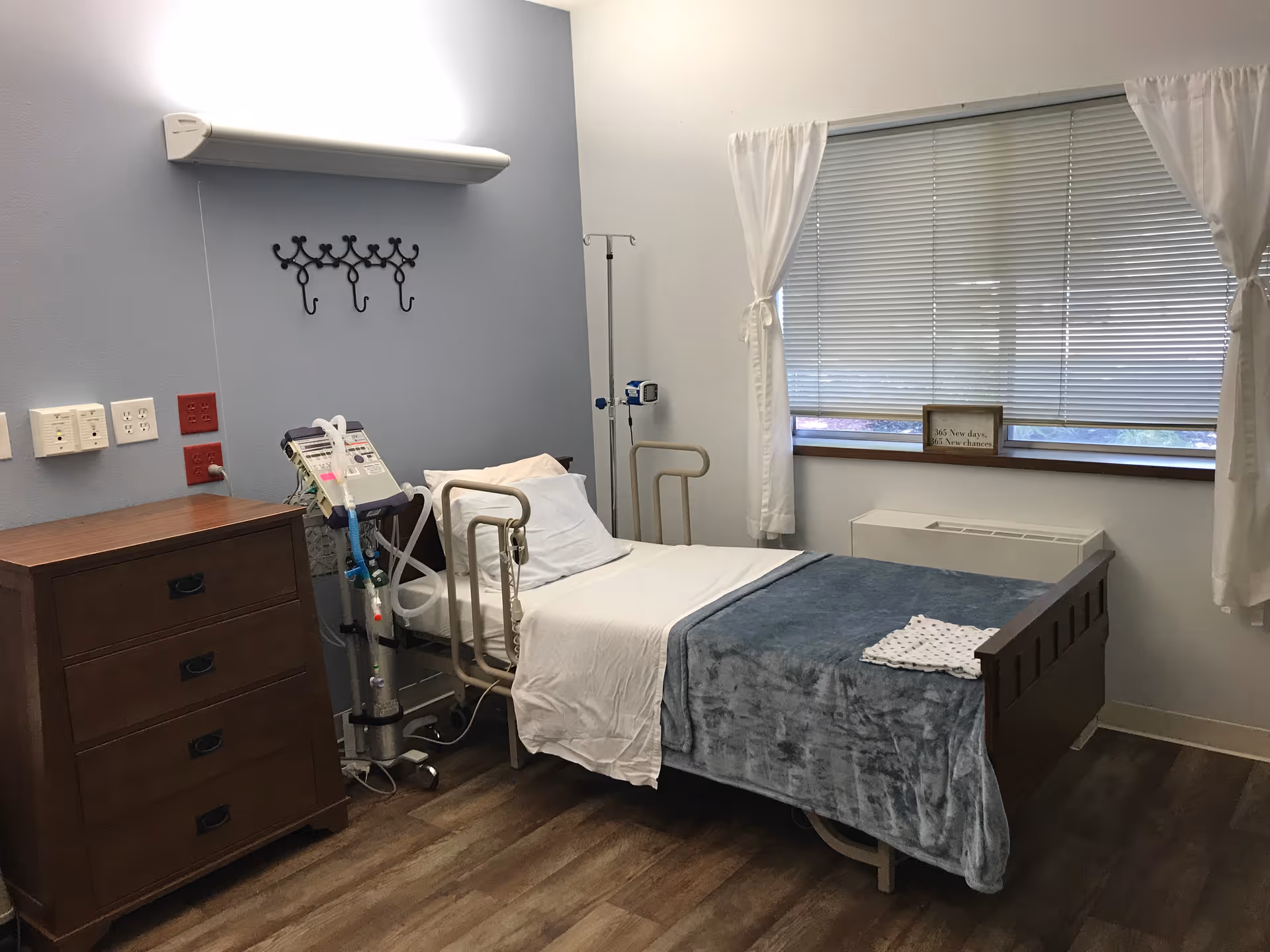 Patient room with a single hospital bed with rails, IV pole and monitoring equipment, a wooden dresser, and a window with blinds.