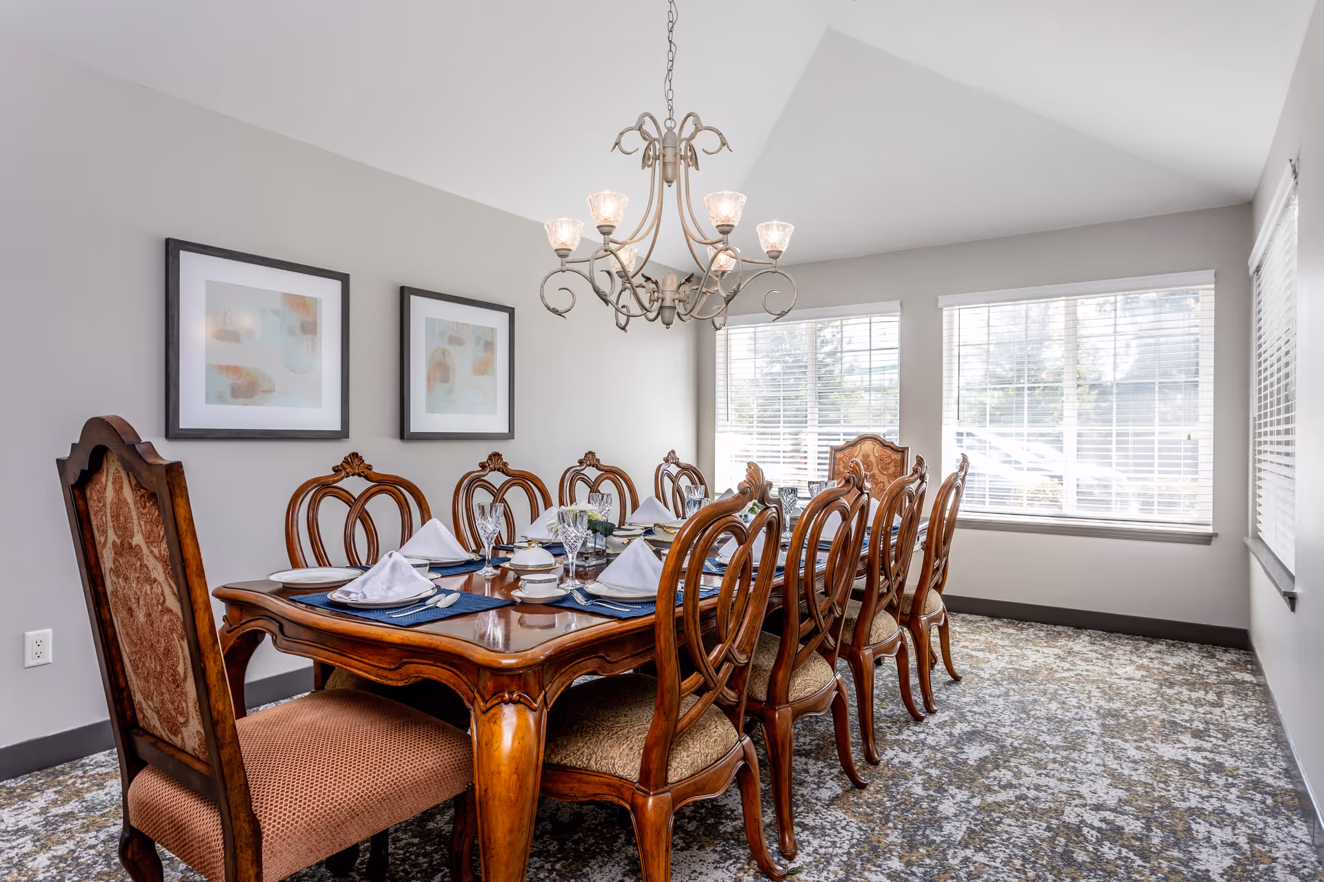 Bright dining room with an ornate wooden table set for a meal, matching carved chairs, a chandelier, and large windows.