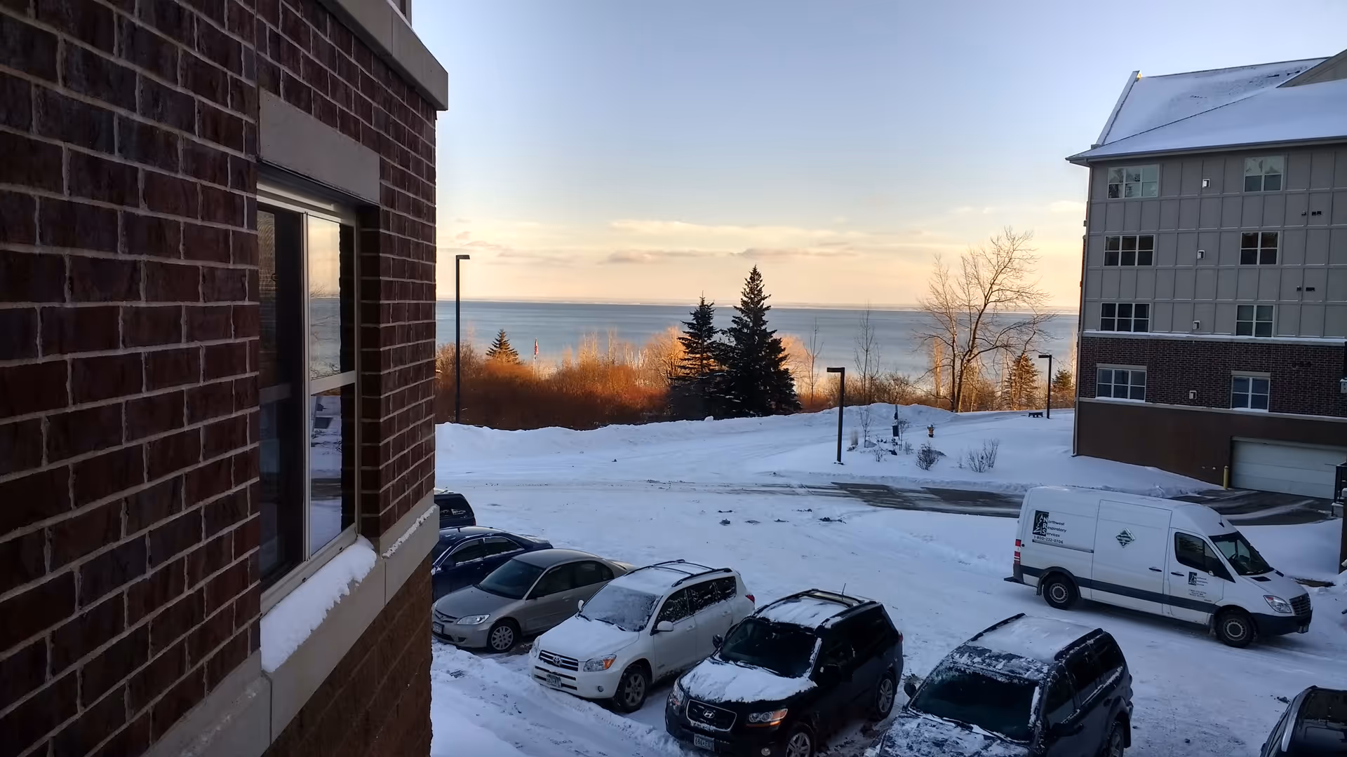 Snow-covered parking lot outside brick buildings with a distant lake horizon.